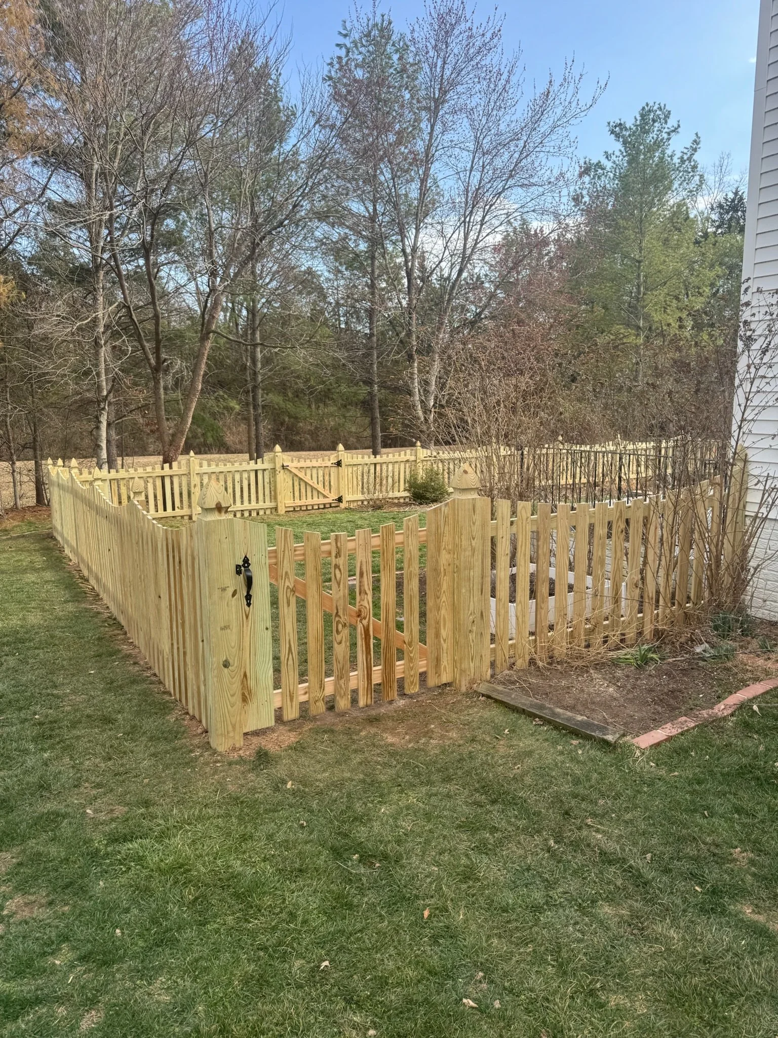 A newly built wooden fence with a gate enclosing a small yard, with trees and a house with white siding in the background.