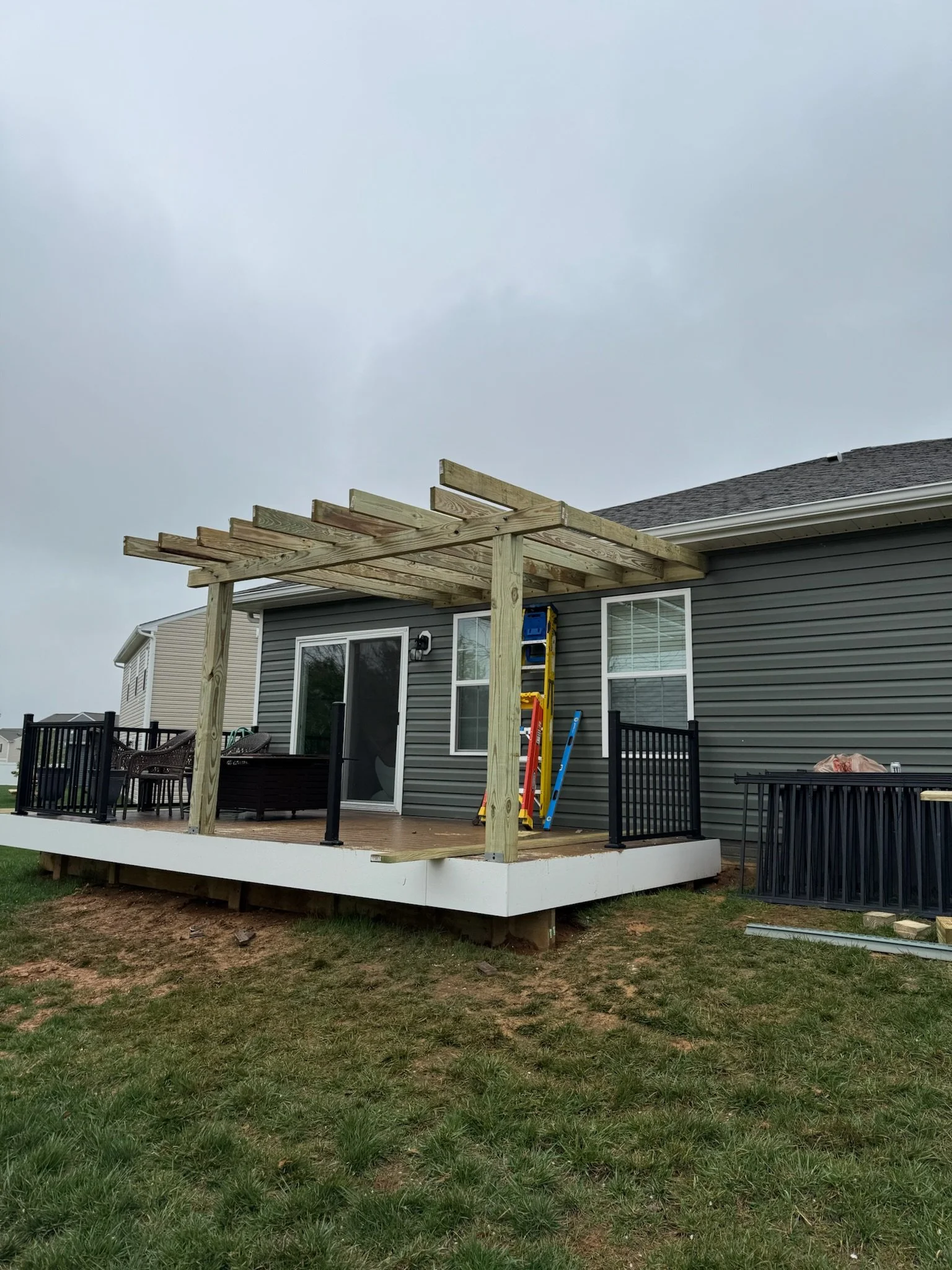 A backyard deck with black railings under construction, attached to a gray house with a sliding glass door and window, and a wooden pergola frame being built over the deck, with construction tools and supplies nearby.