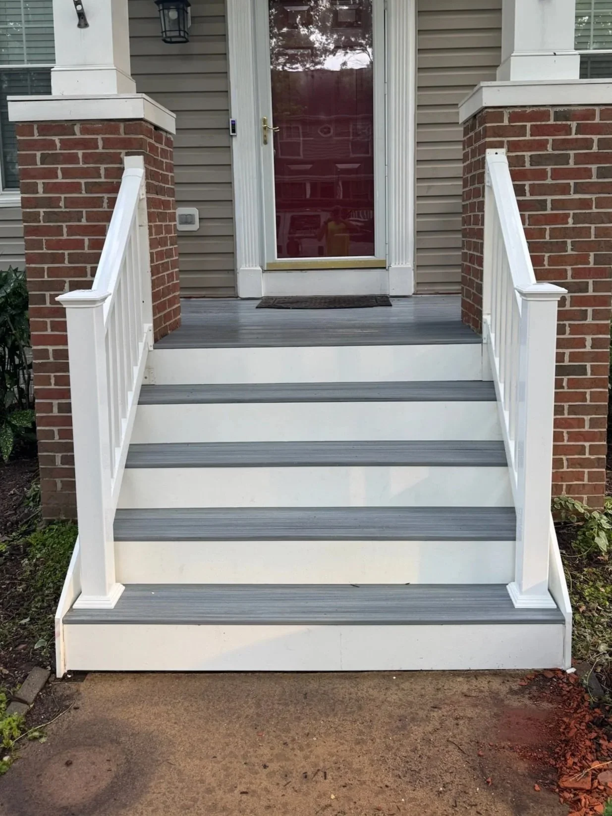 Front porch of a house with a white staircase, brick pillars, and a red front door with a glass panel. There are bushes on both sides of the stairs and a small potted plant on the right. The house number is 434113.