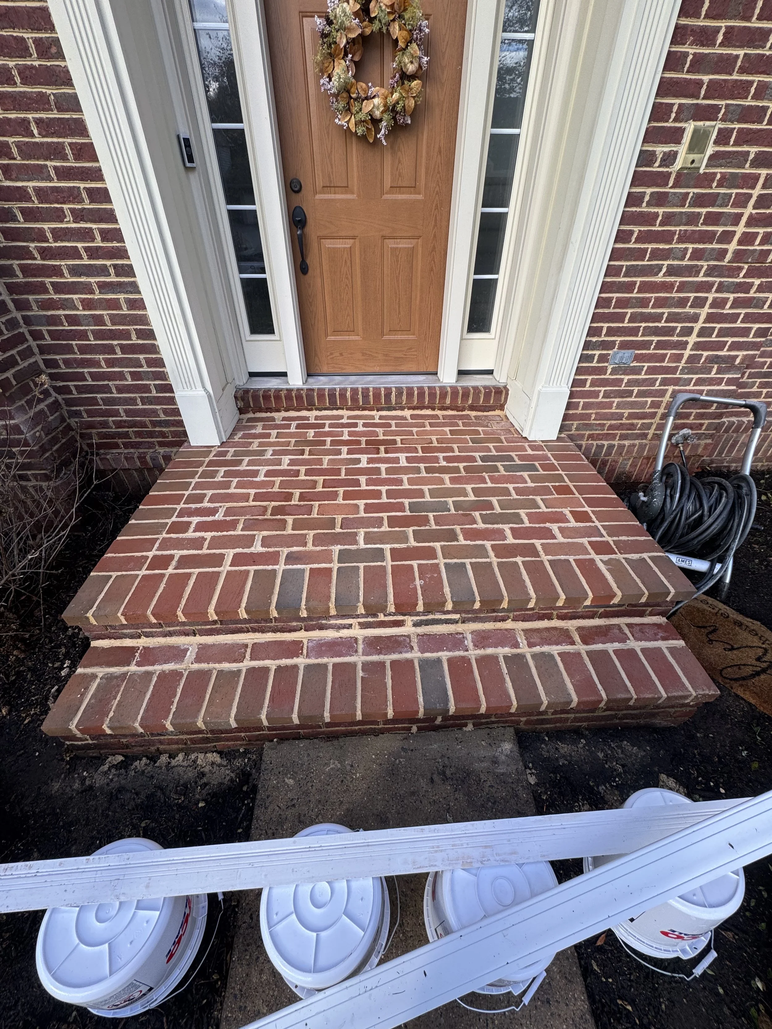 Red brick porch with three steps leading to a wooden front door decorated with a floral wreath, flanked by tall narrow windows. White plastic buckets and a length of white plastic pipe rest on the ground in front of the porch.