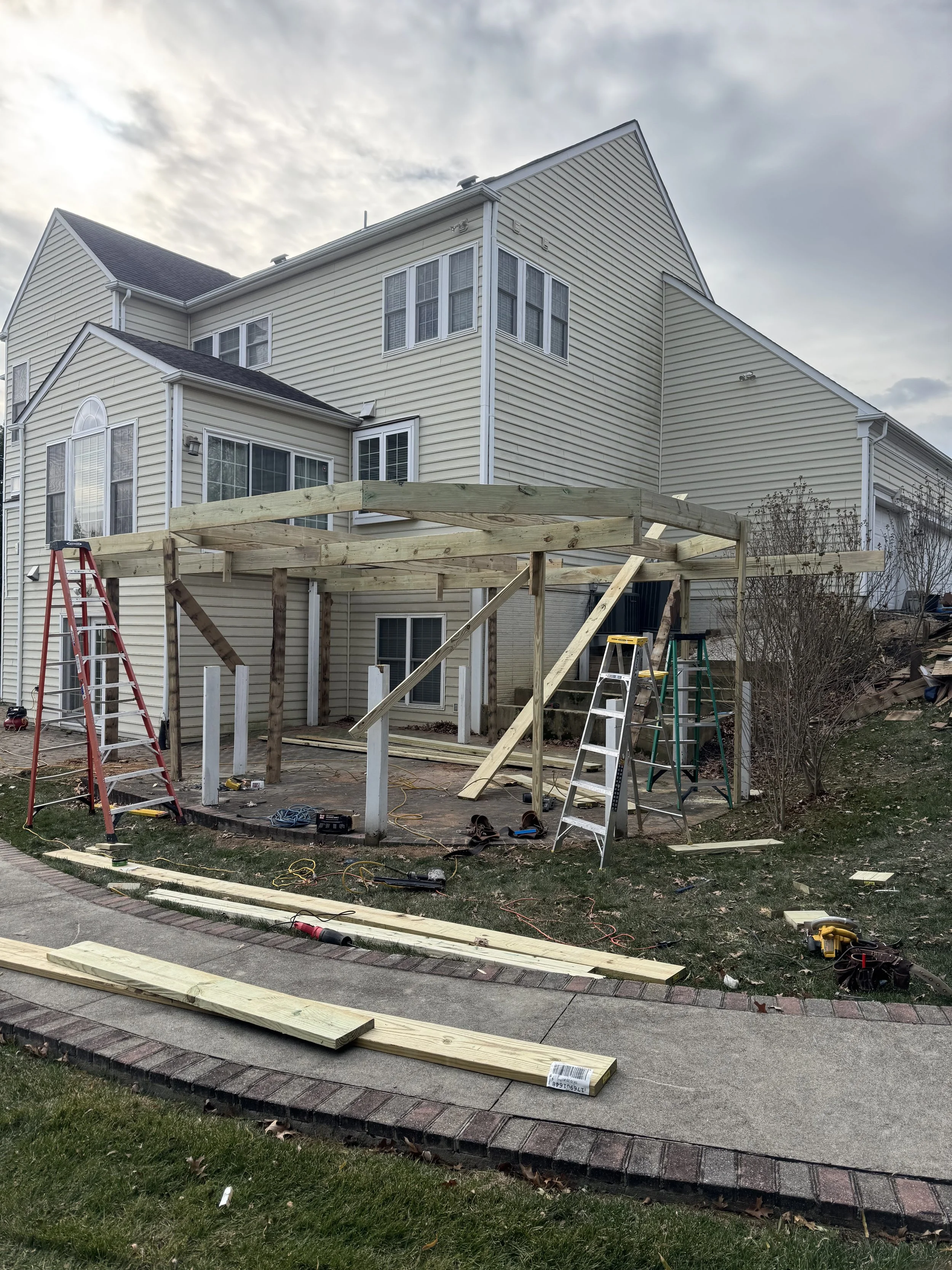 A house under construction with wooden framework for a deck, construction tools, ladders, and building materials scattered around the yard.