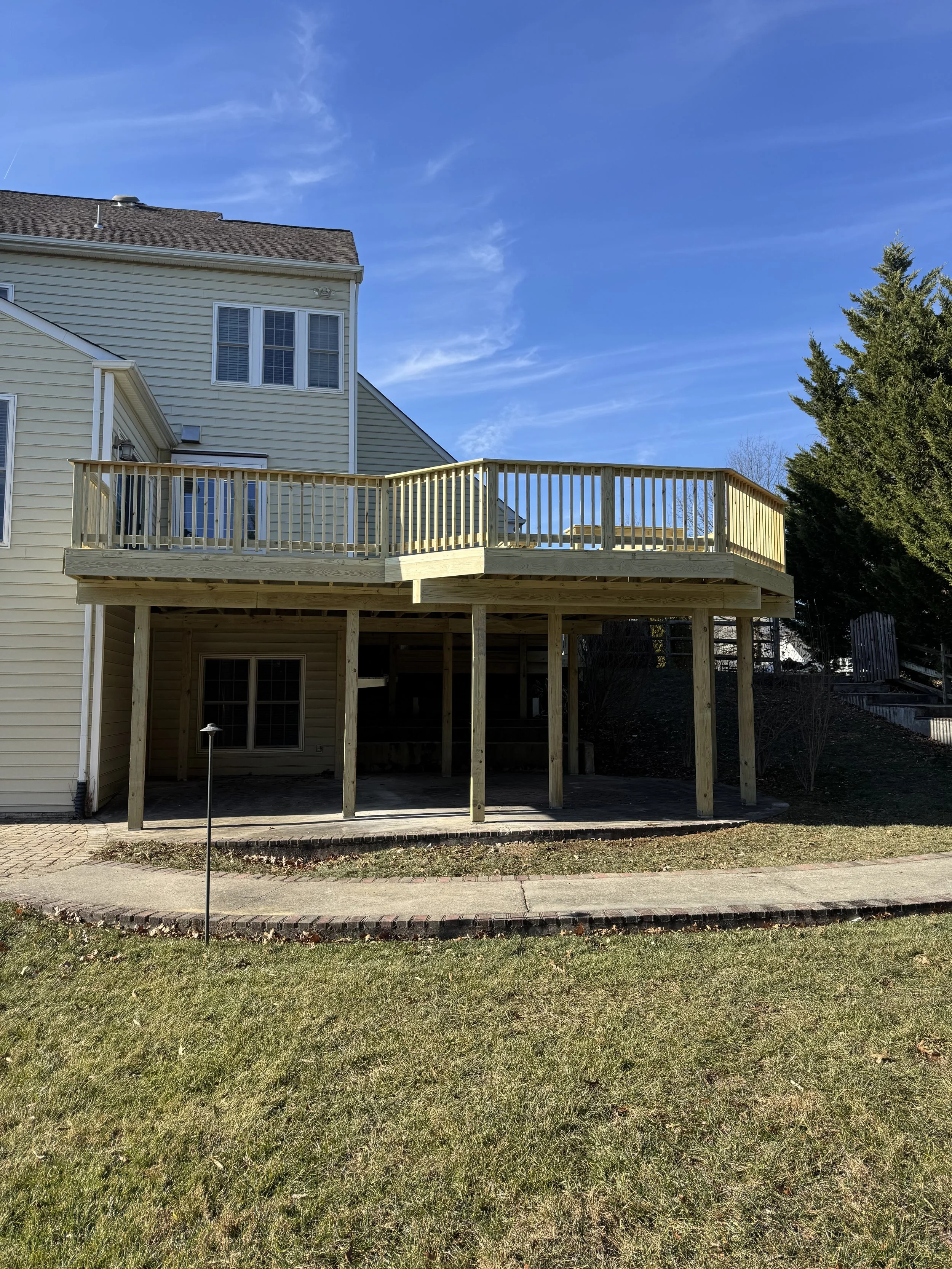 The image shows the exterior of a multi-story house with light yellow siding, and a new wooden deck with railing construction extending from the back of the house. The sky is clear with some wispy clouds, and there are trees and a paved walkway in th