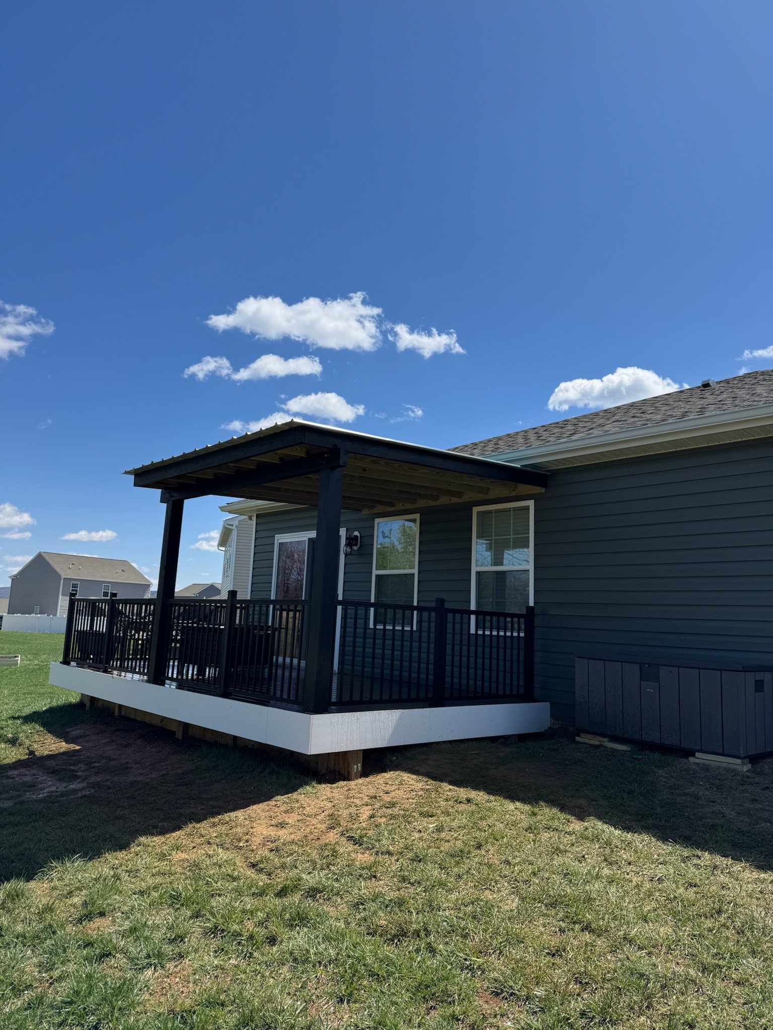 View of a house with a small backyard deck, dark gray siding, and large windows, under a bright blue sky with scattered clouds.