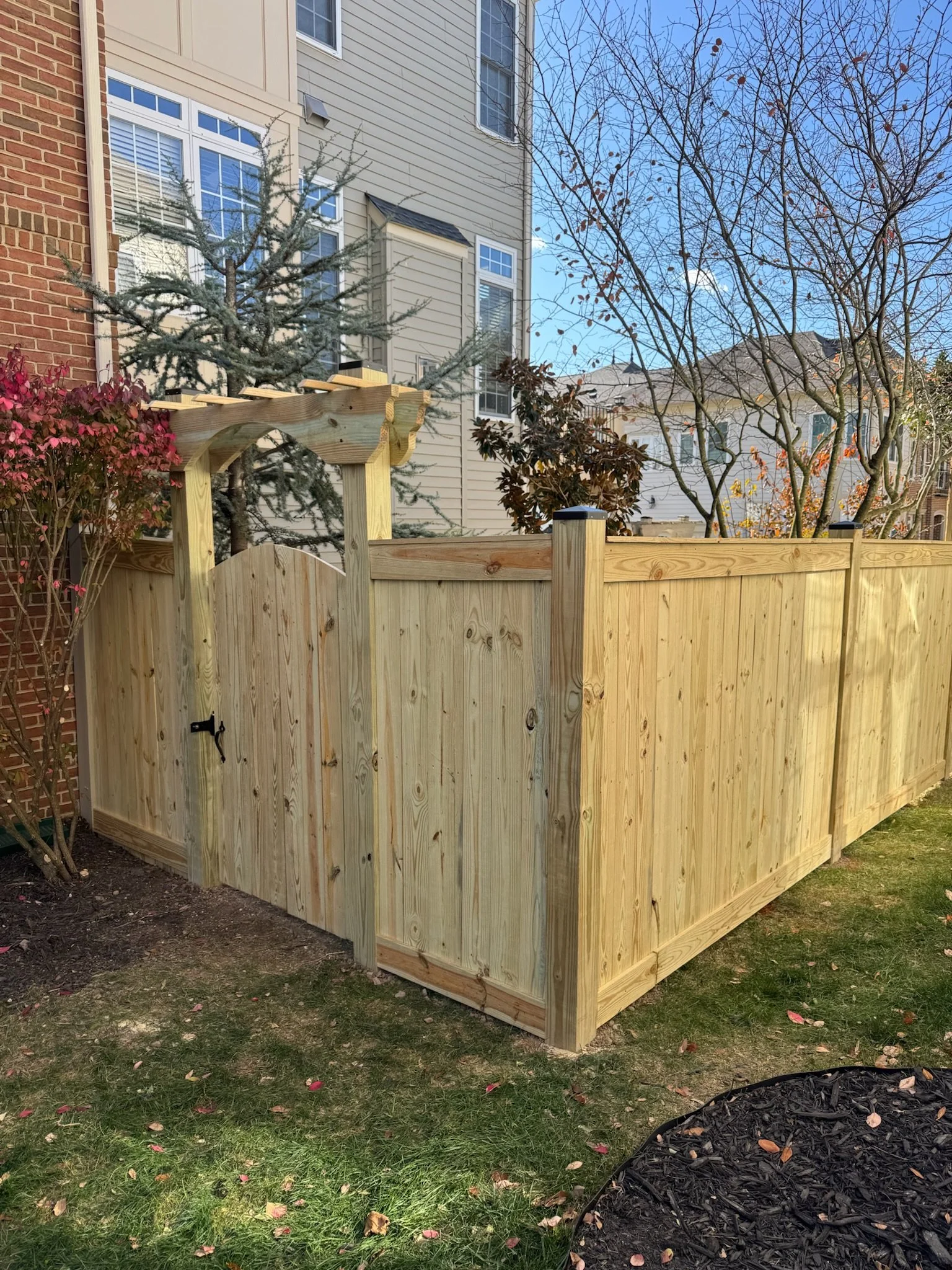 A wooden privacy fence surrounds a backyard with grass, fallen leaves, and mulch in a black plastic garden border. In the background, there are trees with some remaining leaves, neighboring houses, and a partly cloudy blue sky.