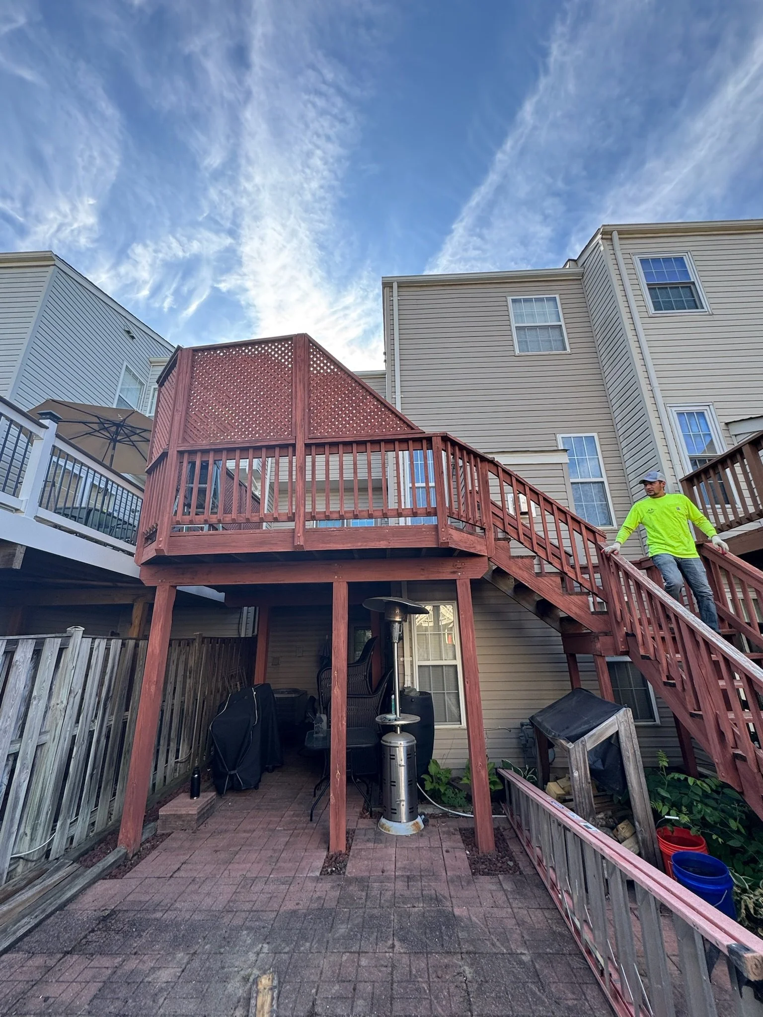 A backyard patio area with a red wooden deck and stairs attached to a beige house with multiple windows. The patio underneath has some patio furniture, an outdoor heater, and other items. A man in a bright yellow shirt and gray pants is walking down 
