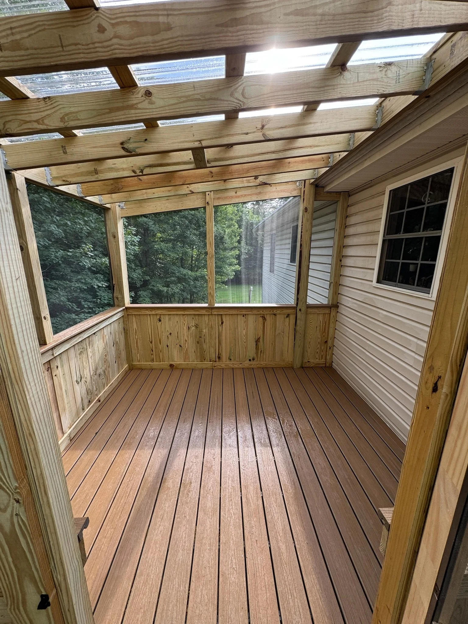 A wooden screened porch attached to a house with vinyl siding, featuring a wood floor and a window, overlooking trees outside.