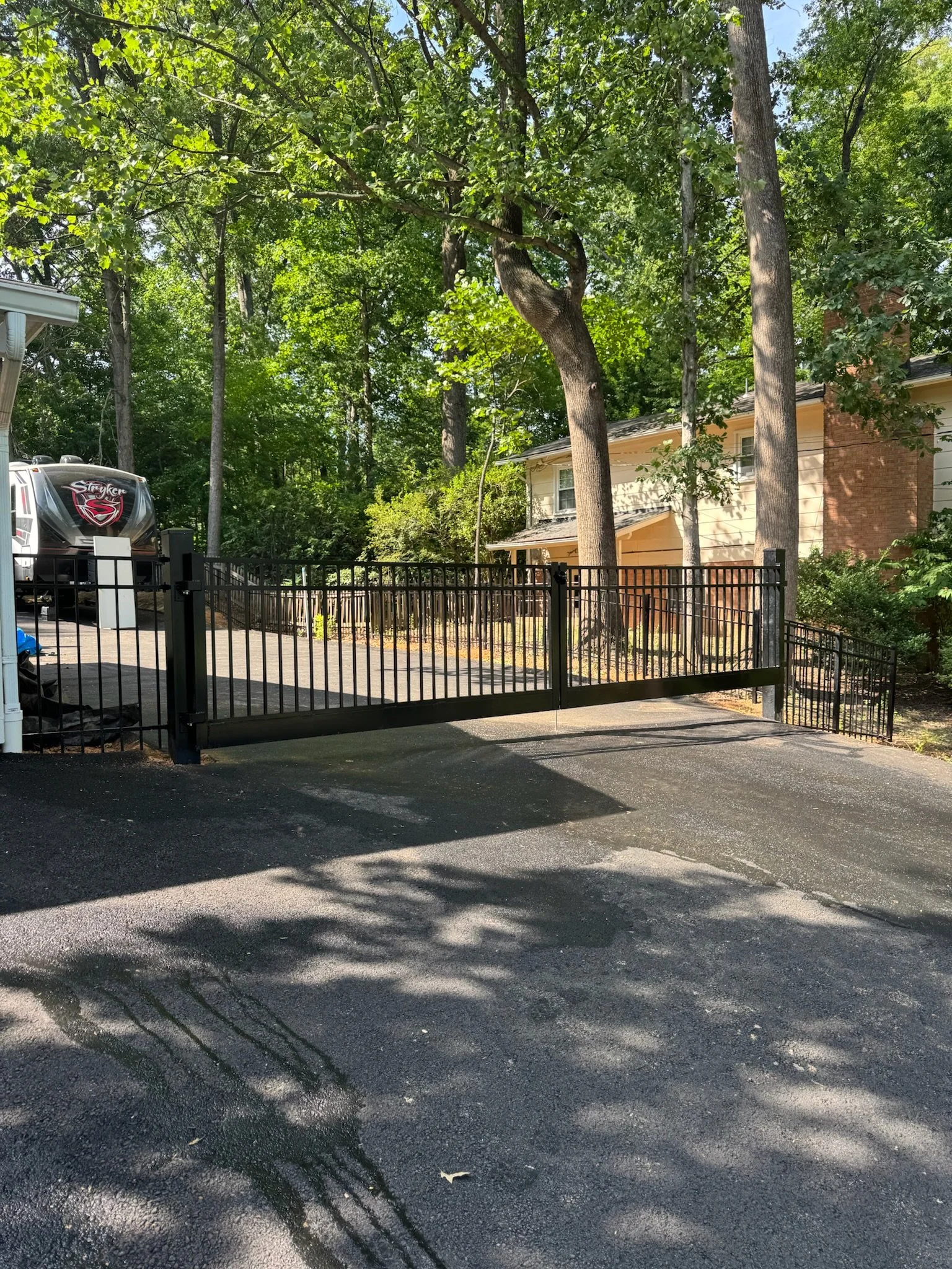 A black metal gate in front of a driveway with a house and trees in the background, and shadows cast on the paved ground.