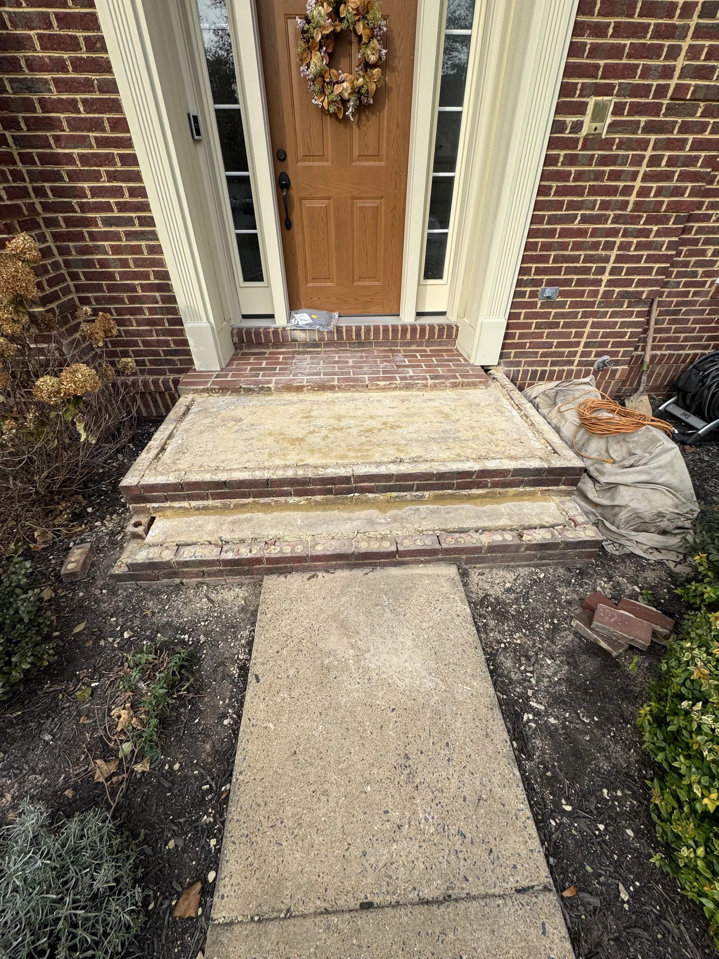 The image shows the front entrance of a house with a wooden door. There are three concrete steps leading up to the door, which is surrounded by brick walls and vertical side windows. A wreath with dried flowers and leaves is hanging on the door. To t