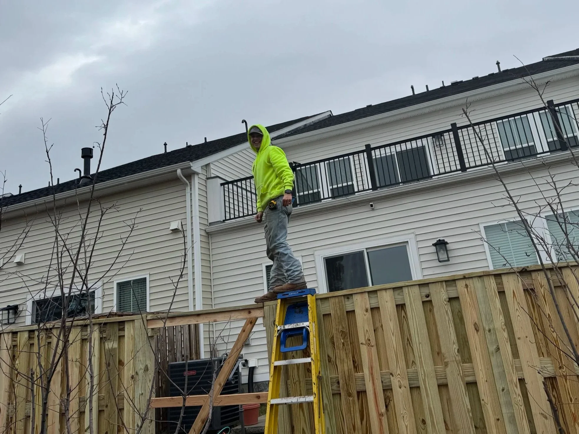 A person wearing a bright yellow hoodie and gray pants standing on a blue and yellow ladder in a backyard, smiling at the camera. The backyard has a wooden fence, some leafless trees, and a two-story house with white siding and black railing balconies under a cloudy sky.