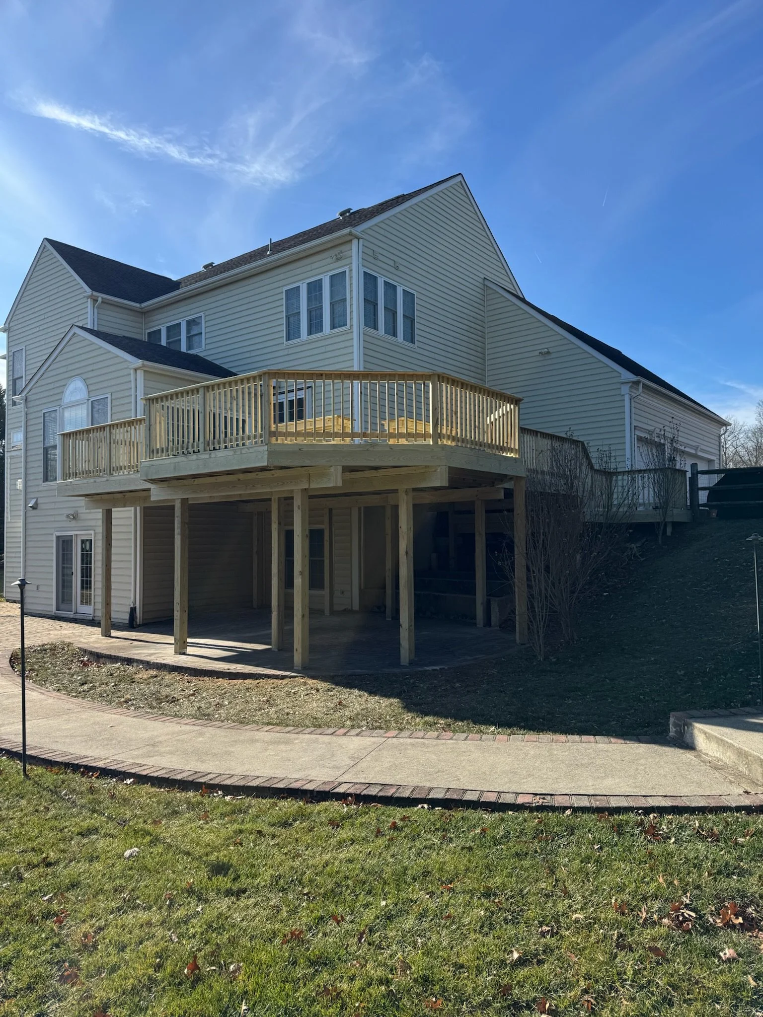 A two-story house with beige siding and white trim; there is a newly built wooden deck outside with no furniture; a grassy yard and a sidewalk are in the foreground, and the sky is clear and blue.