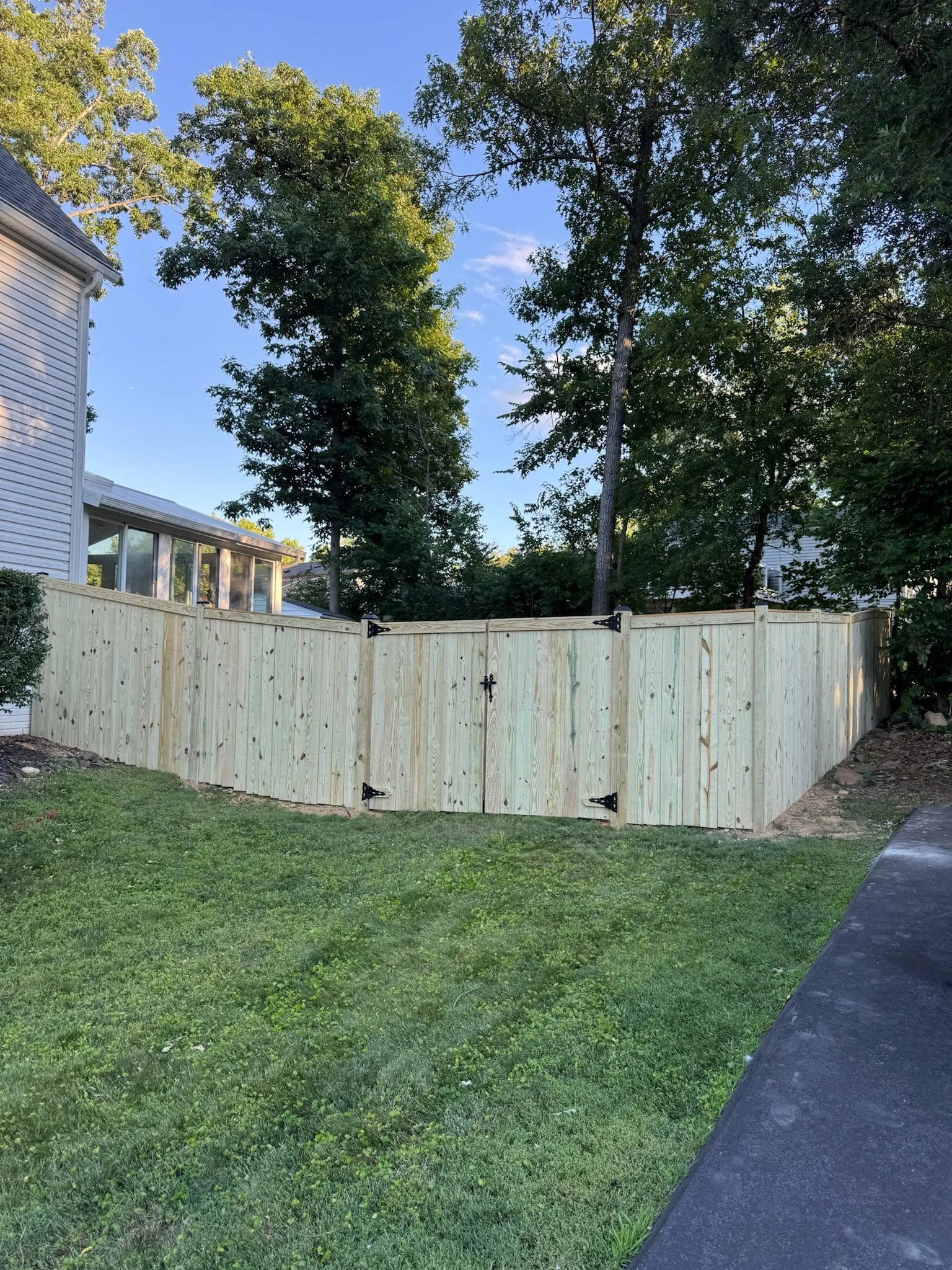 Backyard with a wooden fence, green grass, and trees, with a corner of a house visible on the left side and a paved path on the right.