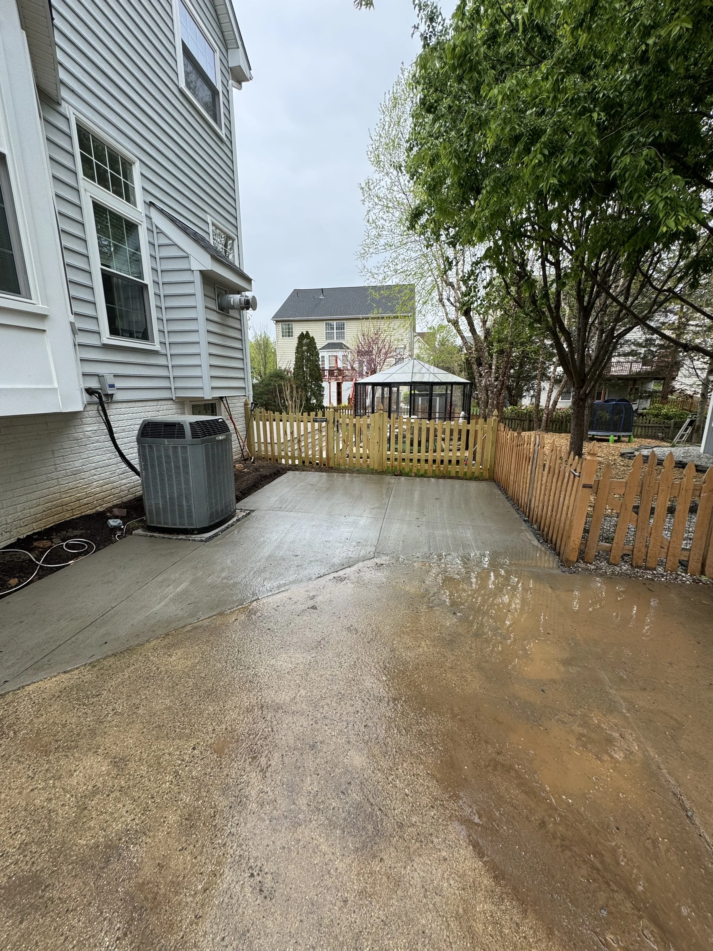 A wet concrete patio area next to a house with siding, a fenced backyard with trees, a gazebo, and neighboring houses in the background.