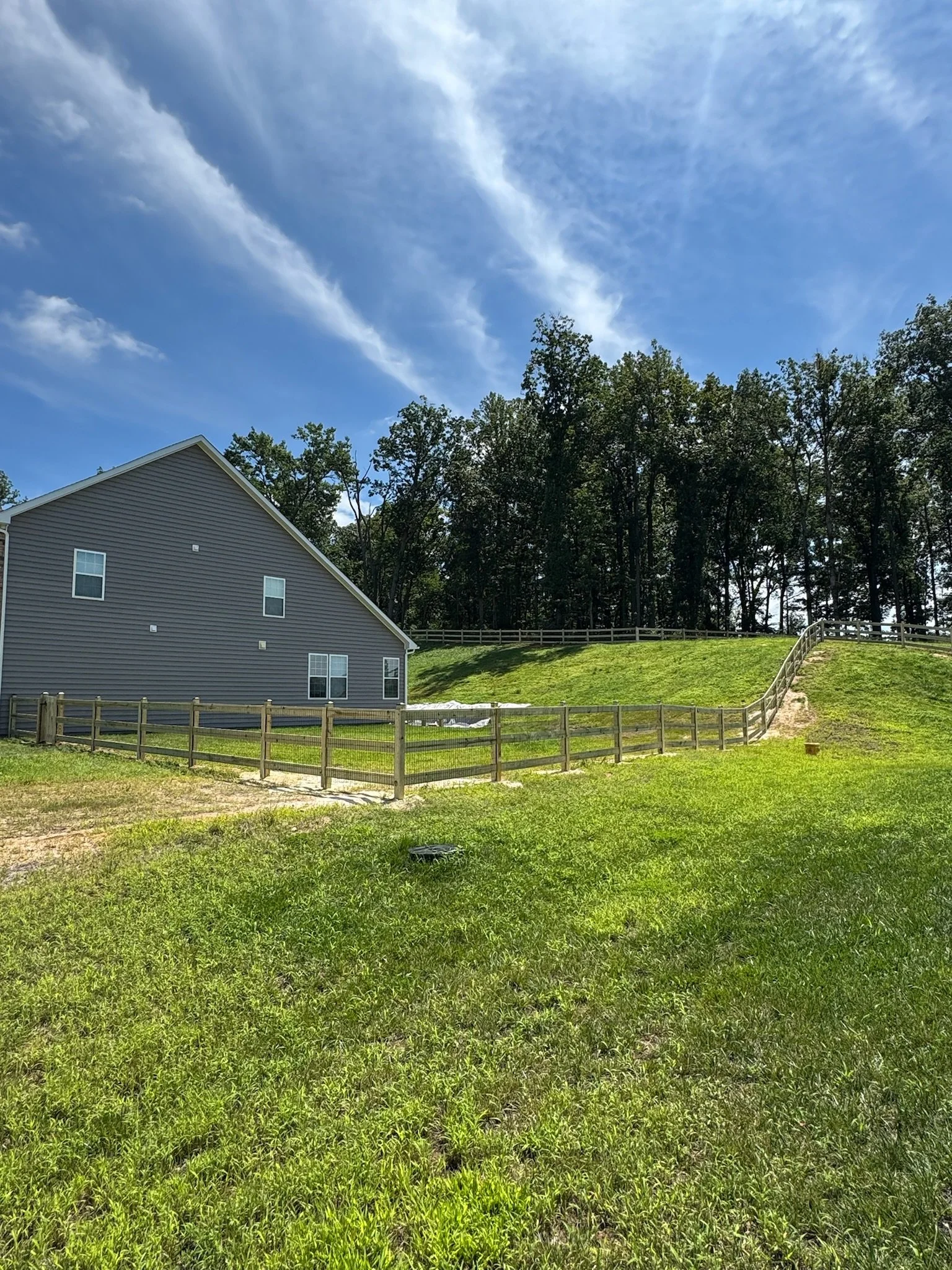 A house with gray siding and white trim is next to a grassy yard enclosed by a wooden fence. Behind the house, there is a hill with a fence on top and a dense line of tall trees. The sky overhead is mostly clear with a few wispy clouds.