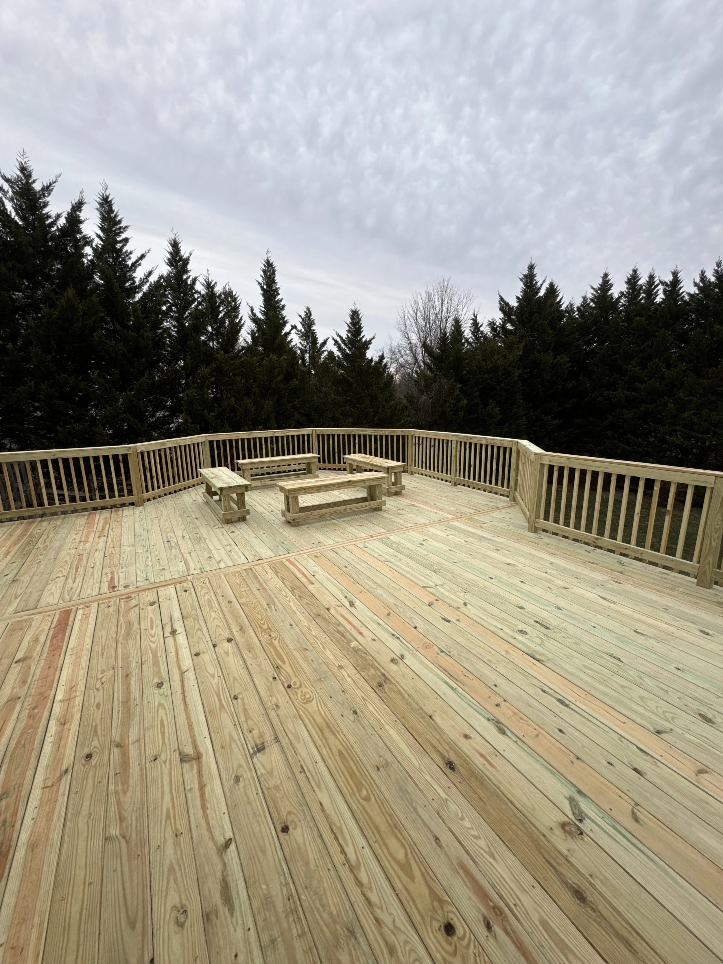 A wooden deck with benches, surrounded by tall evergreen trees under a cloudy sky.