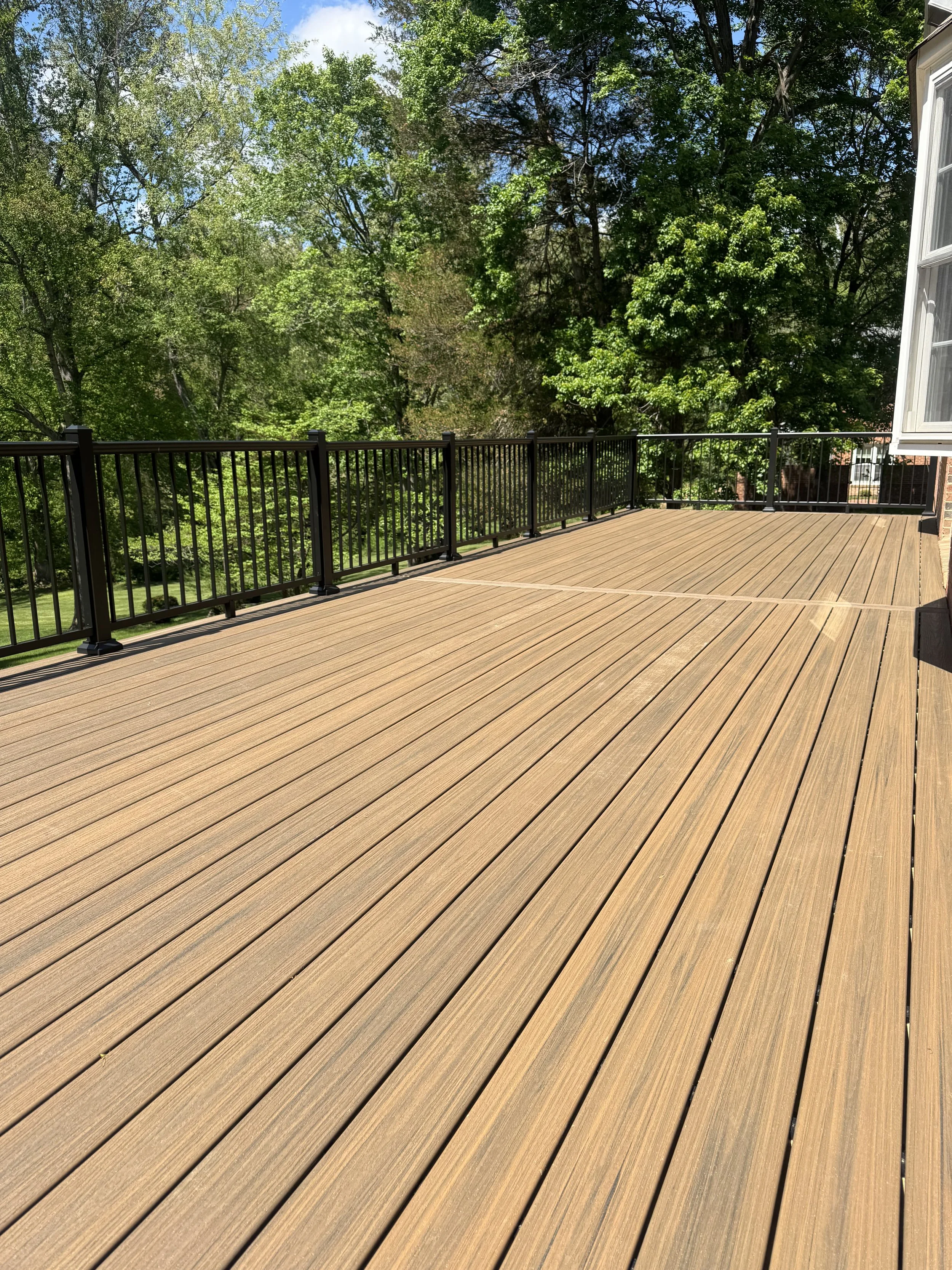 An outdoor wooden deck with black metal railing, surrounded by lush green trees and a blue sky in the background.