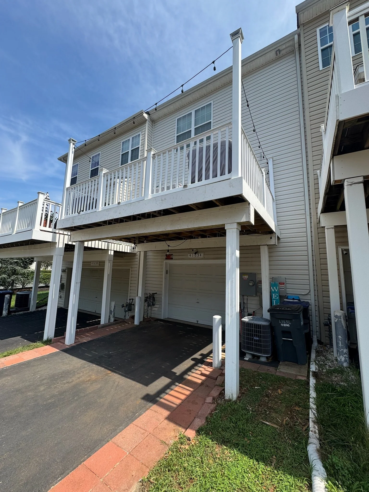 Rear view of a multi-story residential building with a white deck and railing, laundry covered with a tarp, visible garage doors on the ground floor, and a clear blue sky with some clouds.