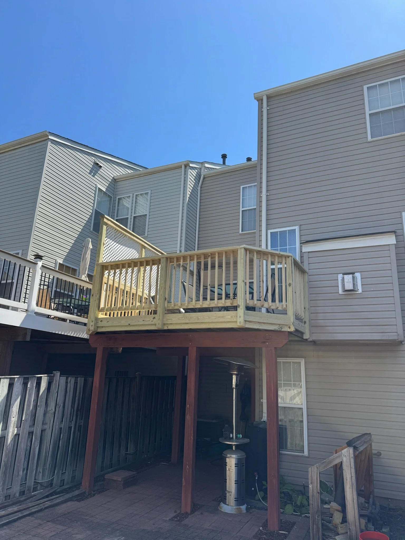 Backyard patio area with an unfinished wooden deck and an outdoor grill, fenced with weathered wood fencing, adjoining a beige house with vinyl siding.