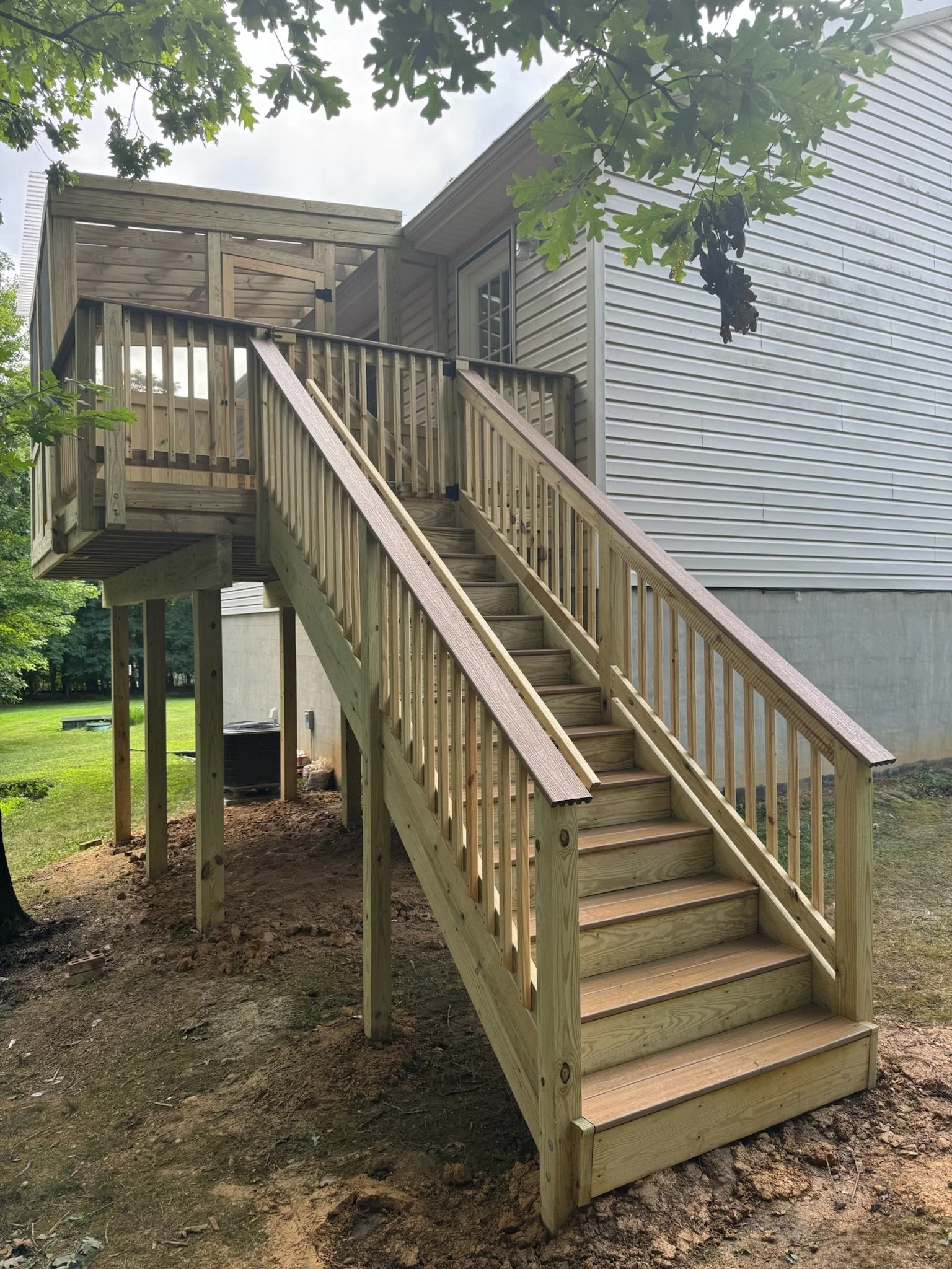 A wooden staircase with a railing leading up to a small balcony or deck attached to the side of a house with white siding. The area surrounding the stairs is bare soil, and there are green trees and grass in the background.