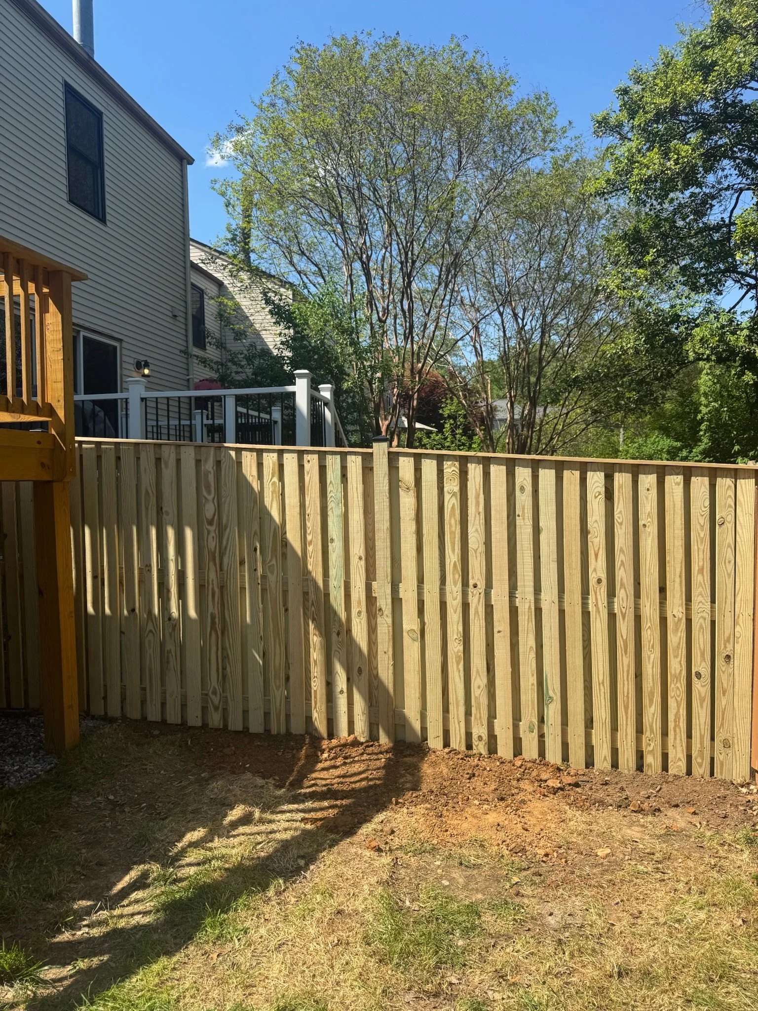 Finished wooden fence in backyard with dirt and grass on ground, house with siding and windows in background, trees with green leaves, and a clear blue sky.