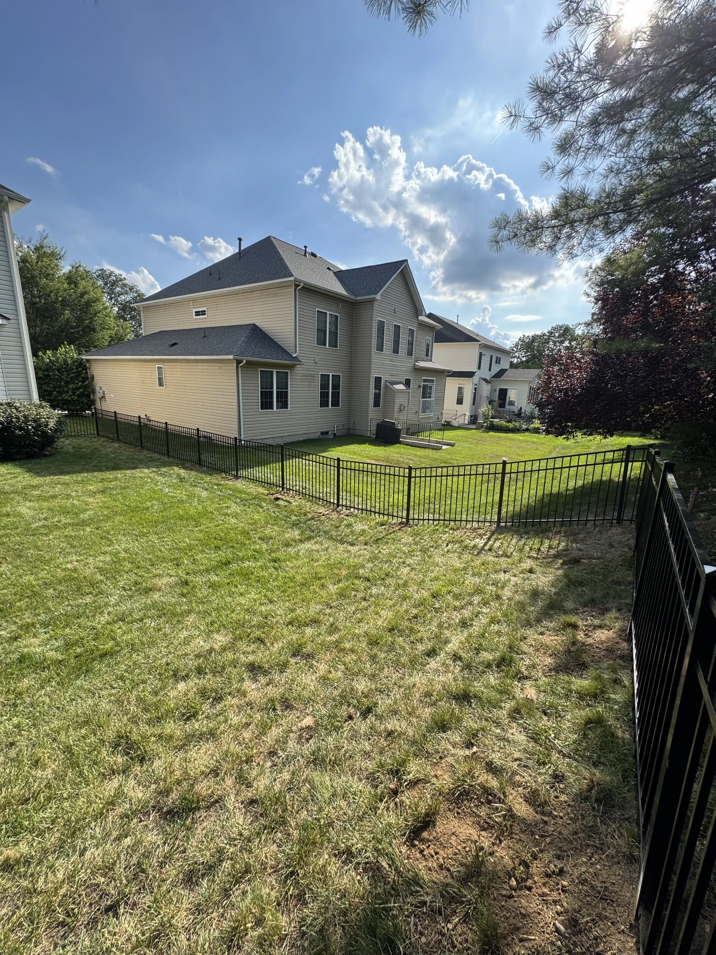 Backyard with green grass, part of a black metal fence, a beige house with multiple windows, and trees with partly cloudy sky.