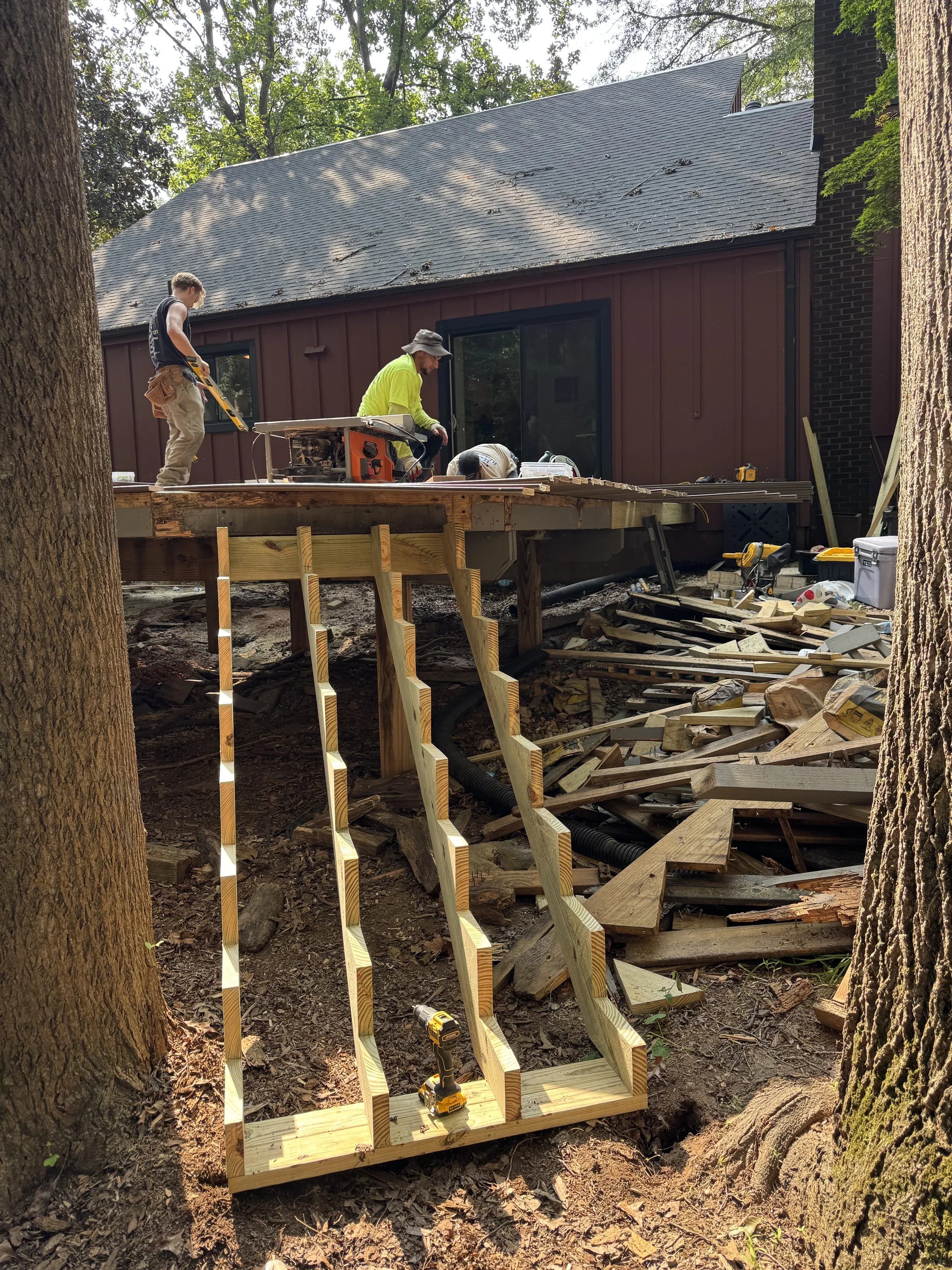Workers building a wooden deck outside a house surrounded by trees.