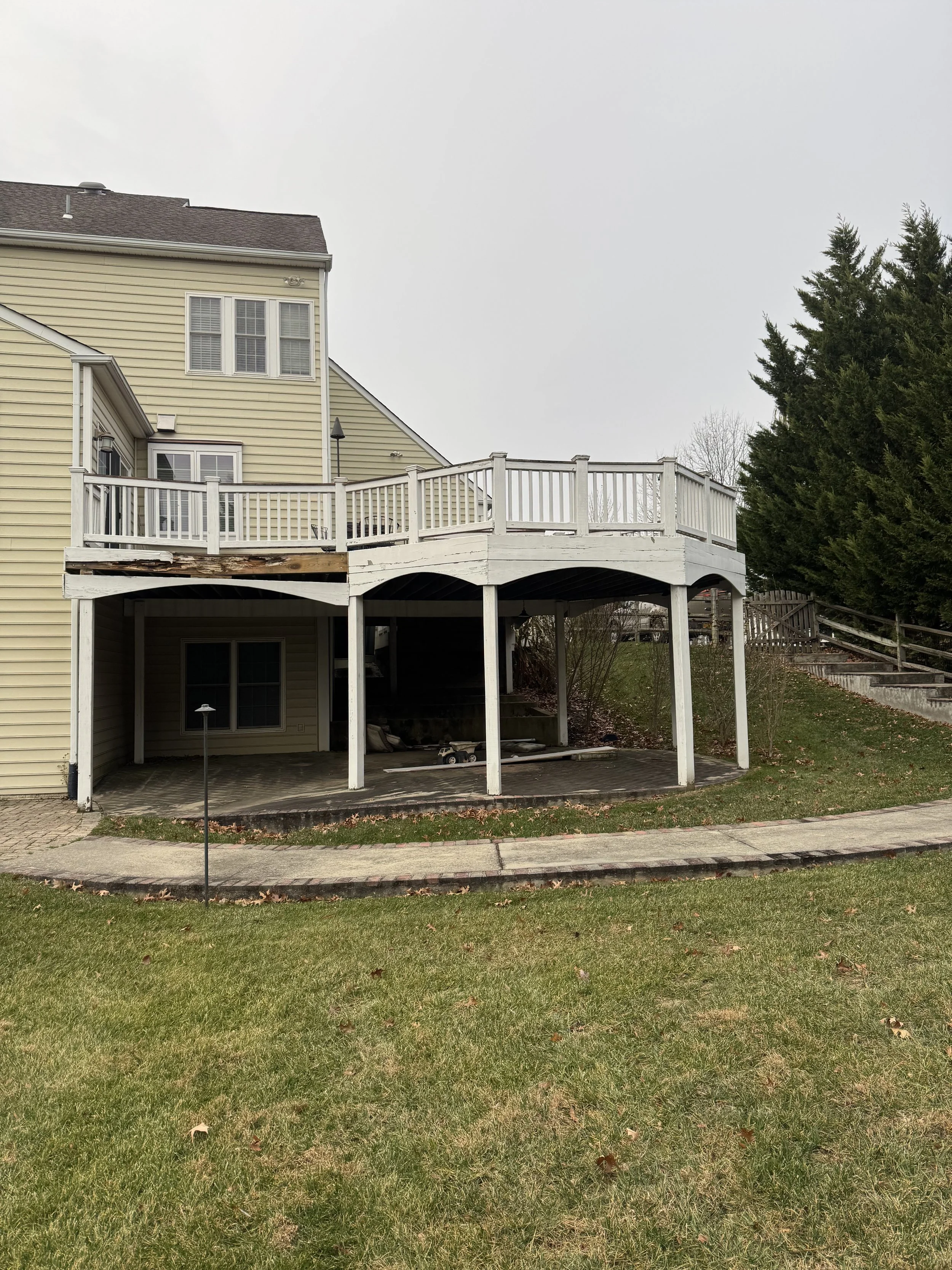 A yellow house with a wooden deck and stairs, a backyard under an overcast sky, a grassy lawn, and green trees.