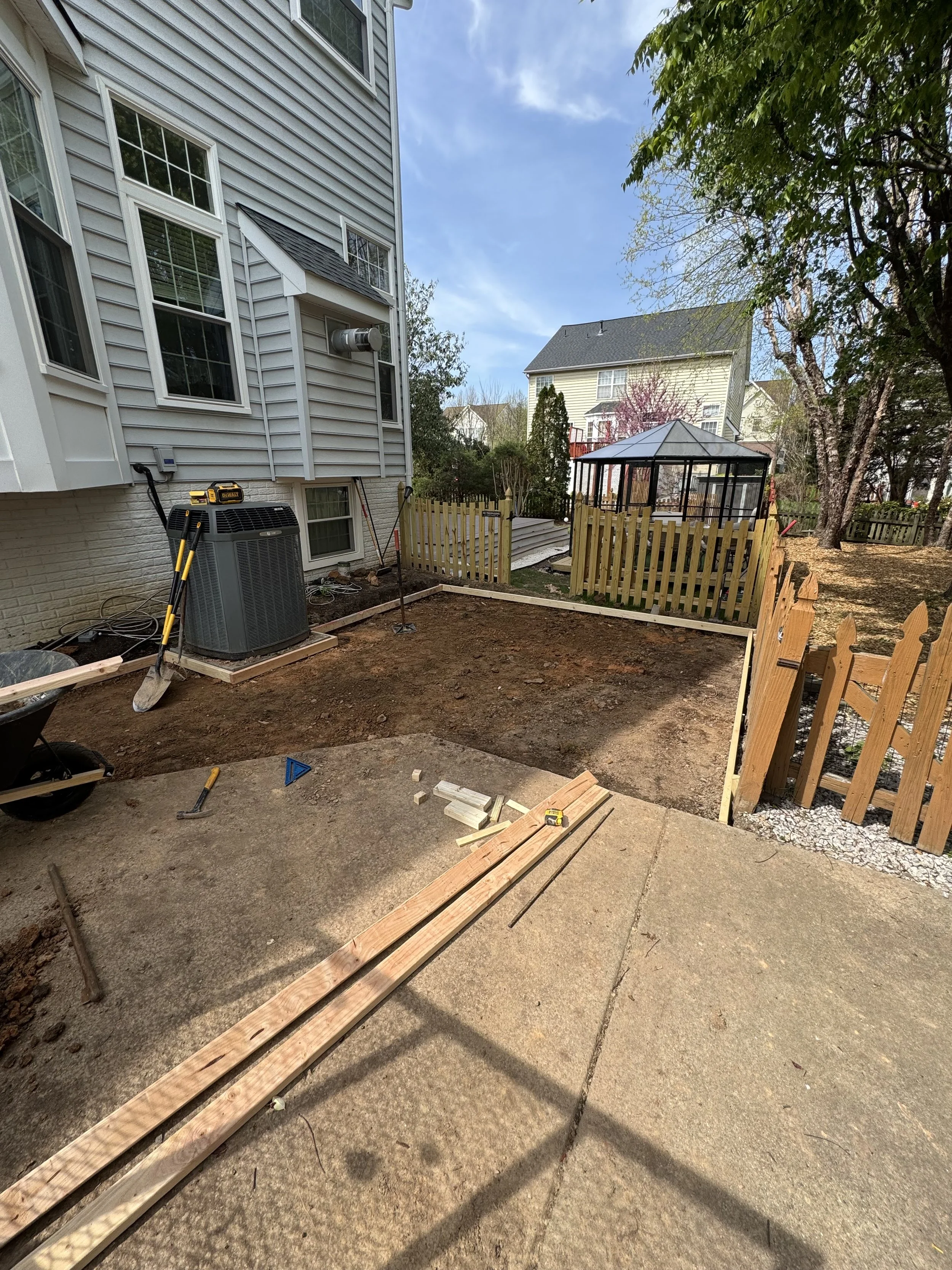 A backyard with construction work in progress, including a wooden fence, a greenhouse, construction tools, and an air conditioning unit.