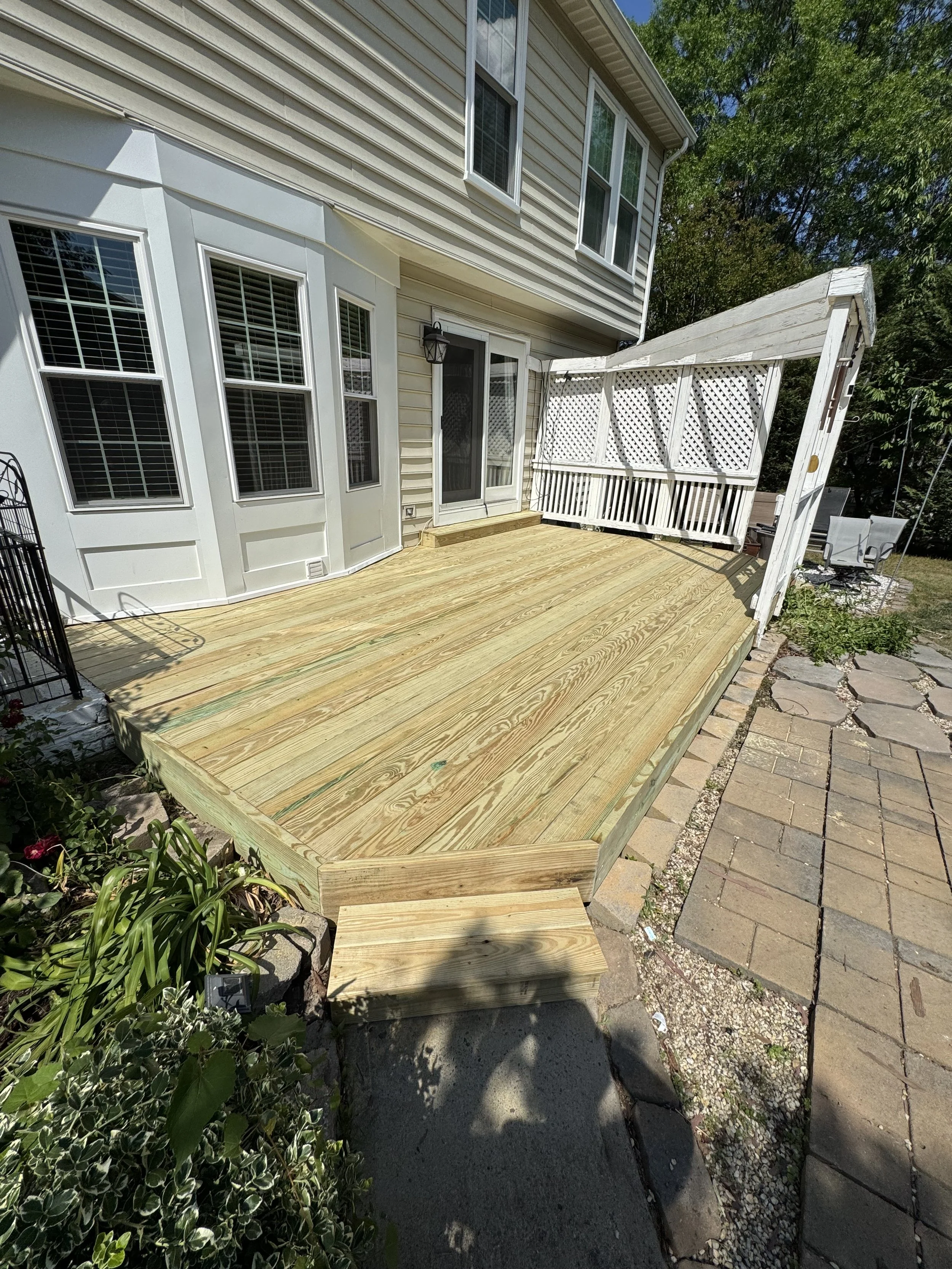 A backyard deck with newly installed wooden flooring, next to a house with beige siding, and surrounded by plants and a brick pathway.