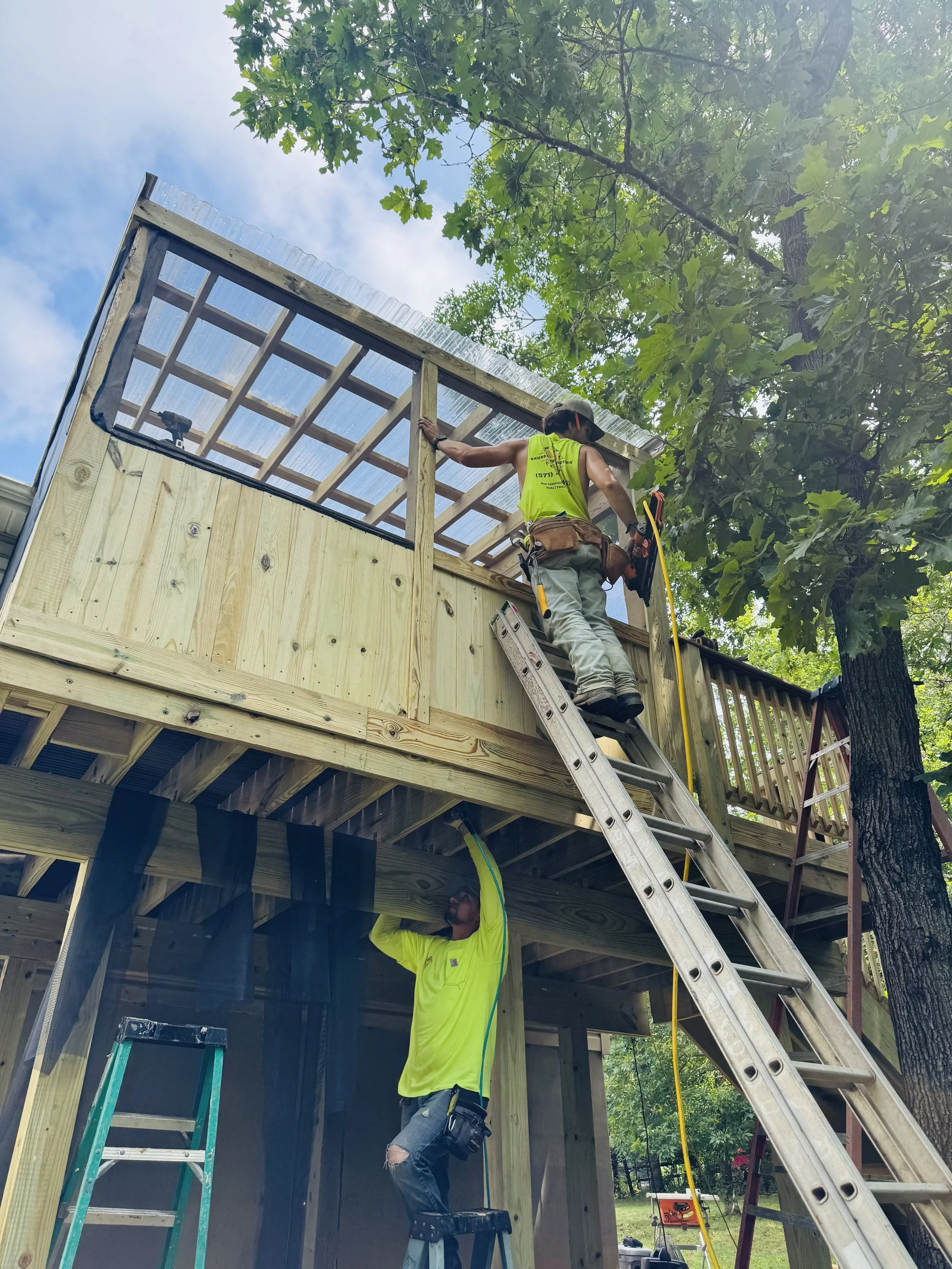 Two construction workers assembling a wooden deck on a house. One is on a ladder, and the other is standing on the ground. The deck has a partly built framed structure with corrugated plastic roofing.