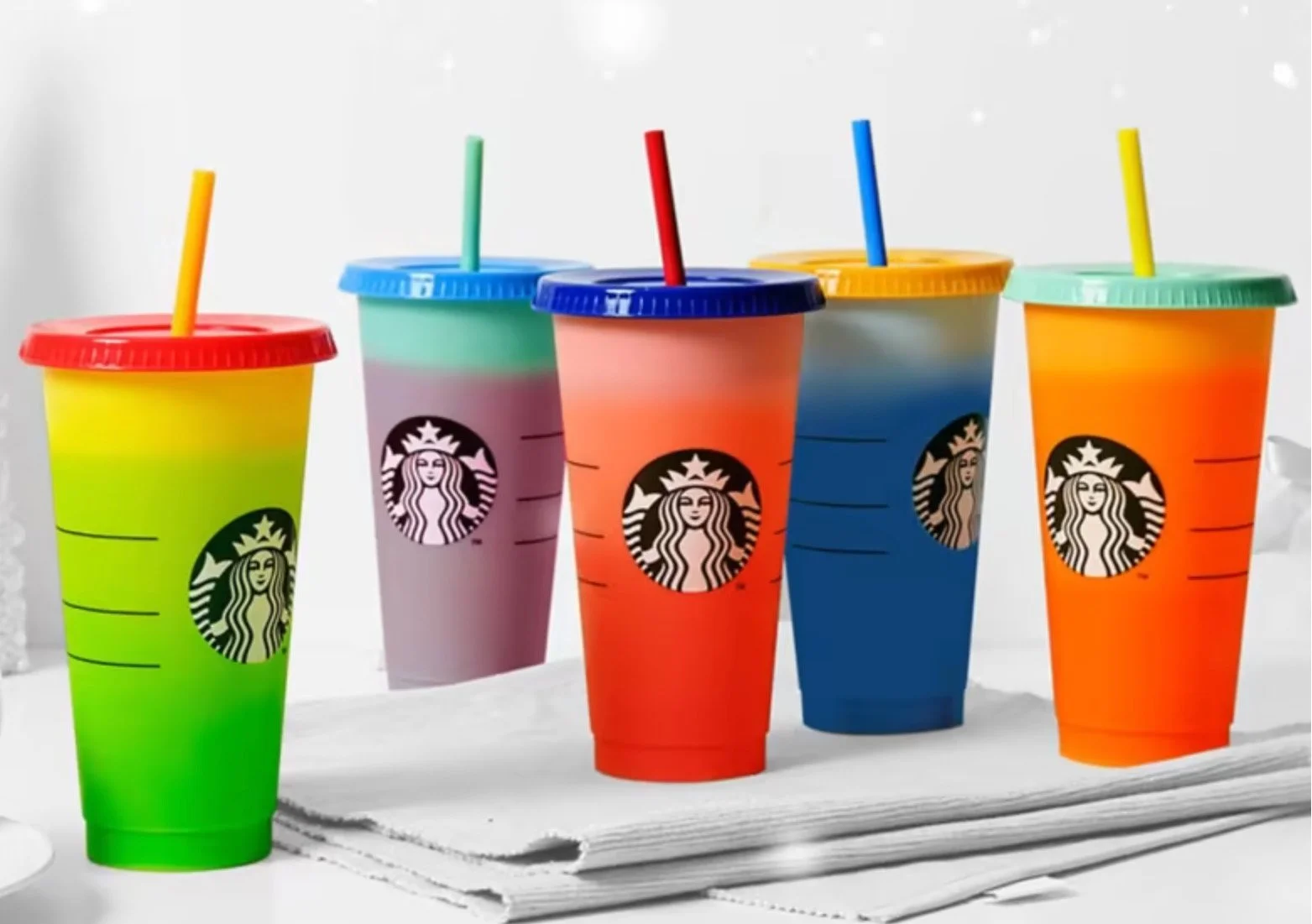 Colorful Starbucks cups with striped designs and straws, arranged on a white surface, against a background with falling snow.