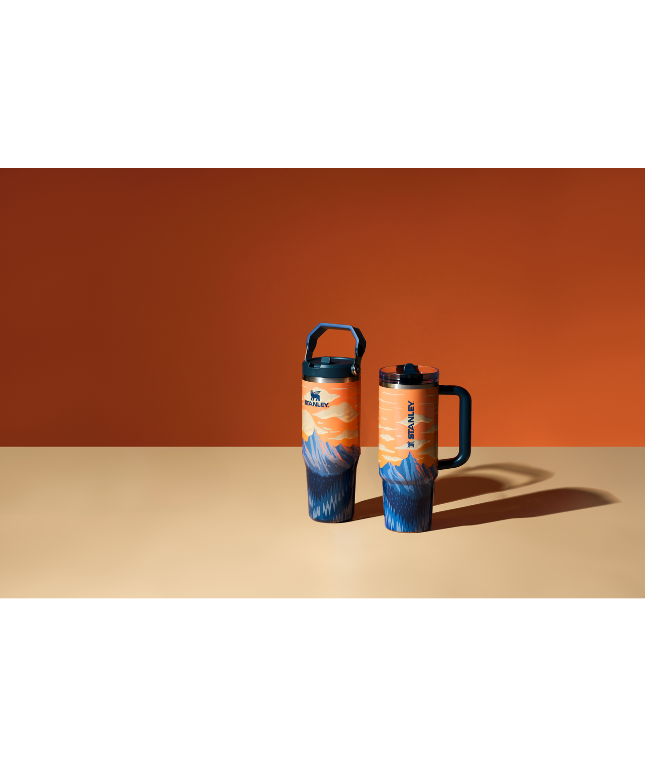 Two Stanley travel mugs with mountain landscape design, one with a handle and the other with a lid, placed on a beige surface with an orange and black background.