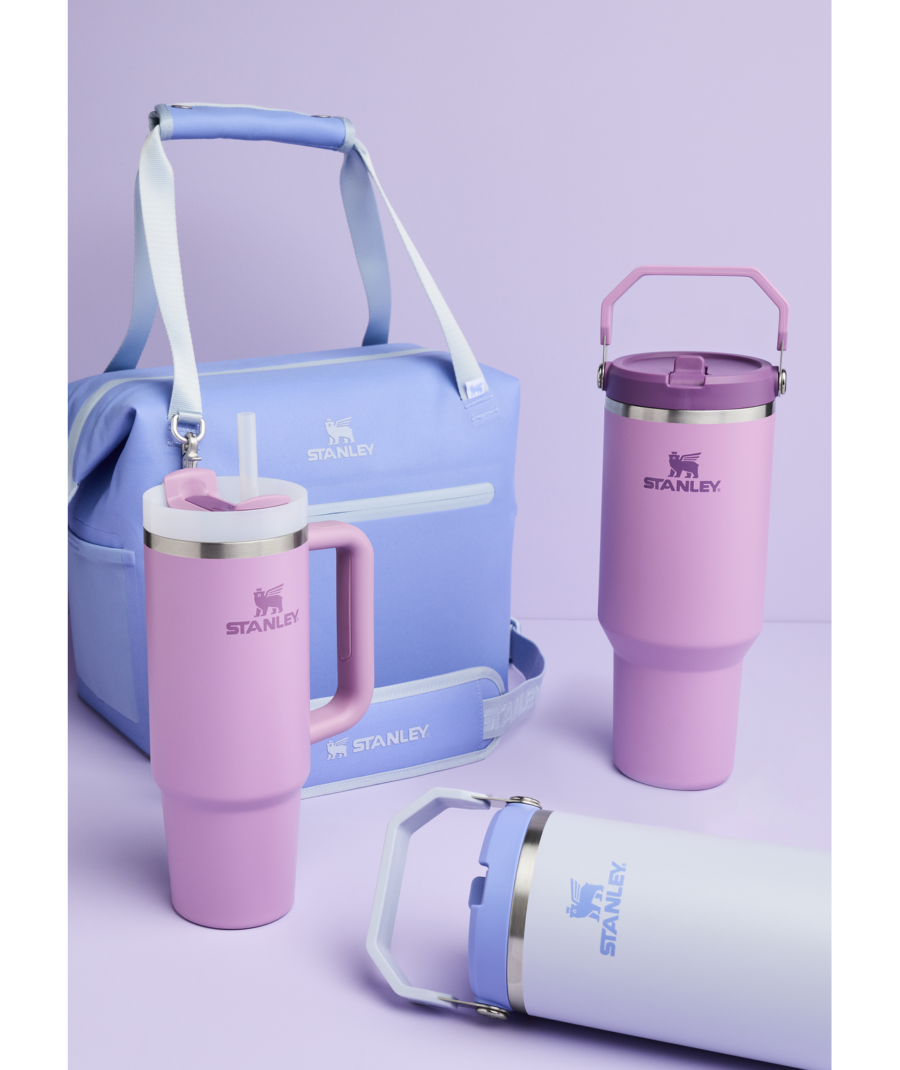 A light purple background with a blue Stanley cooler bag, two pink Stanley insulated tumblers with straws, and a white Stanley flask with blue branding and a handle.