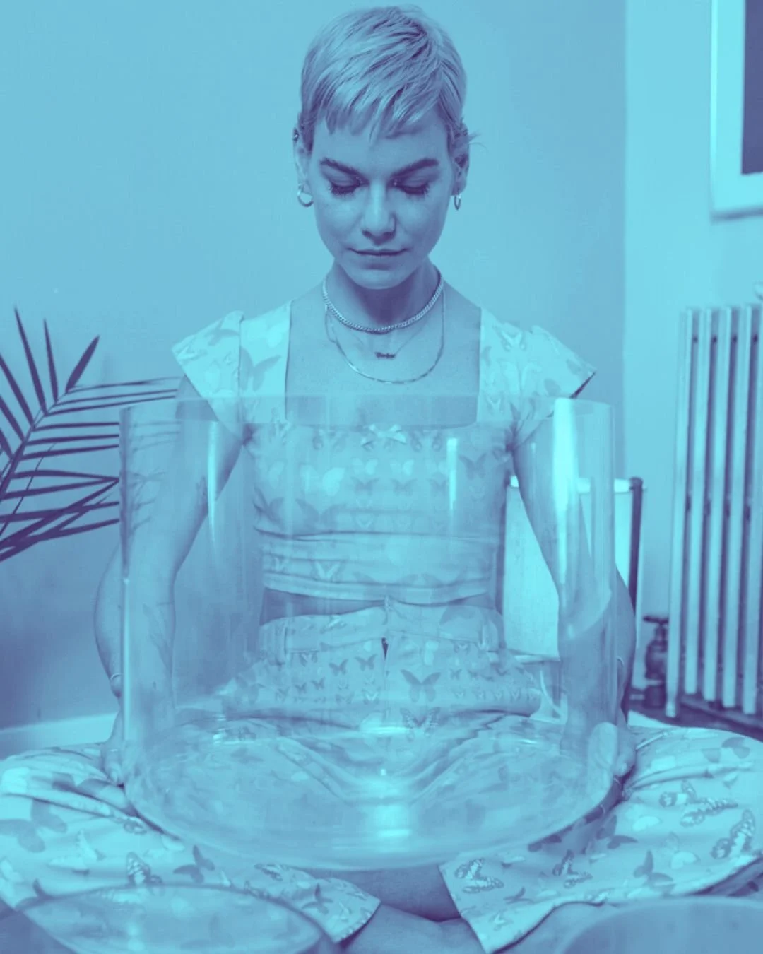A woman with short blond hair looking down at a clear glass bowl in front of her while sitting cross-legged, wearing a butterfly-patterned dress, jewelry, and a silver camisole, in a brightly lit room with plants and a radiator in the background.