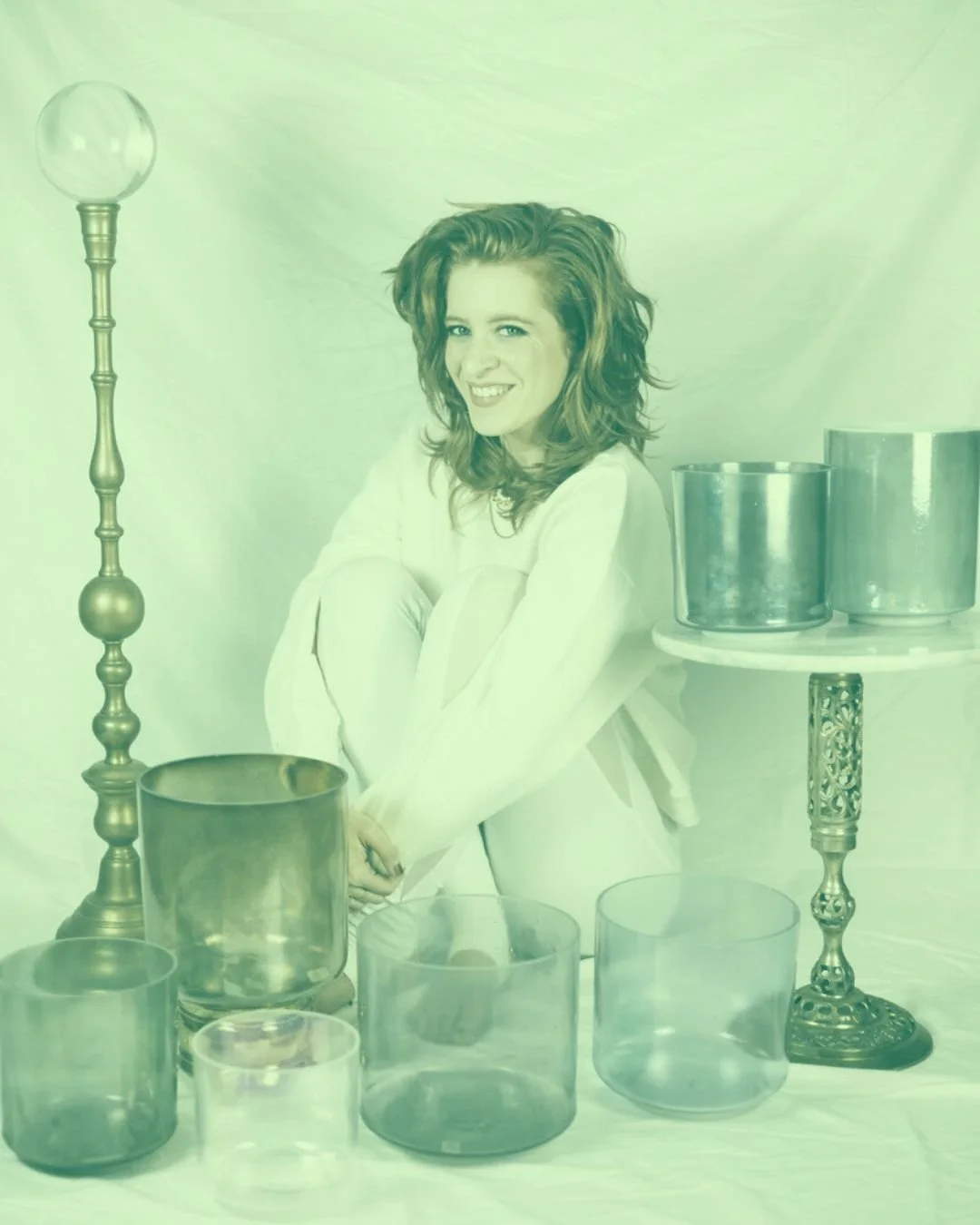 A woman with wavy, red hair wearing a white outfit, sitting among metallic and glass cylindrical objects and standing next to ornate vases and stands, with a plain background.