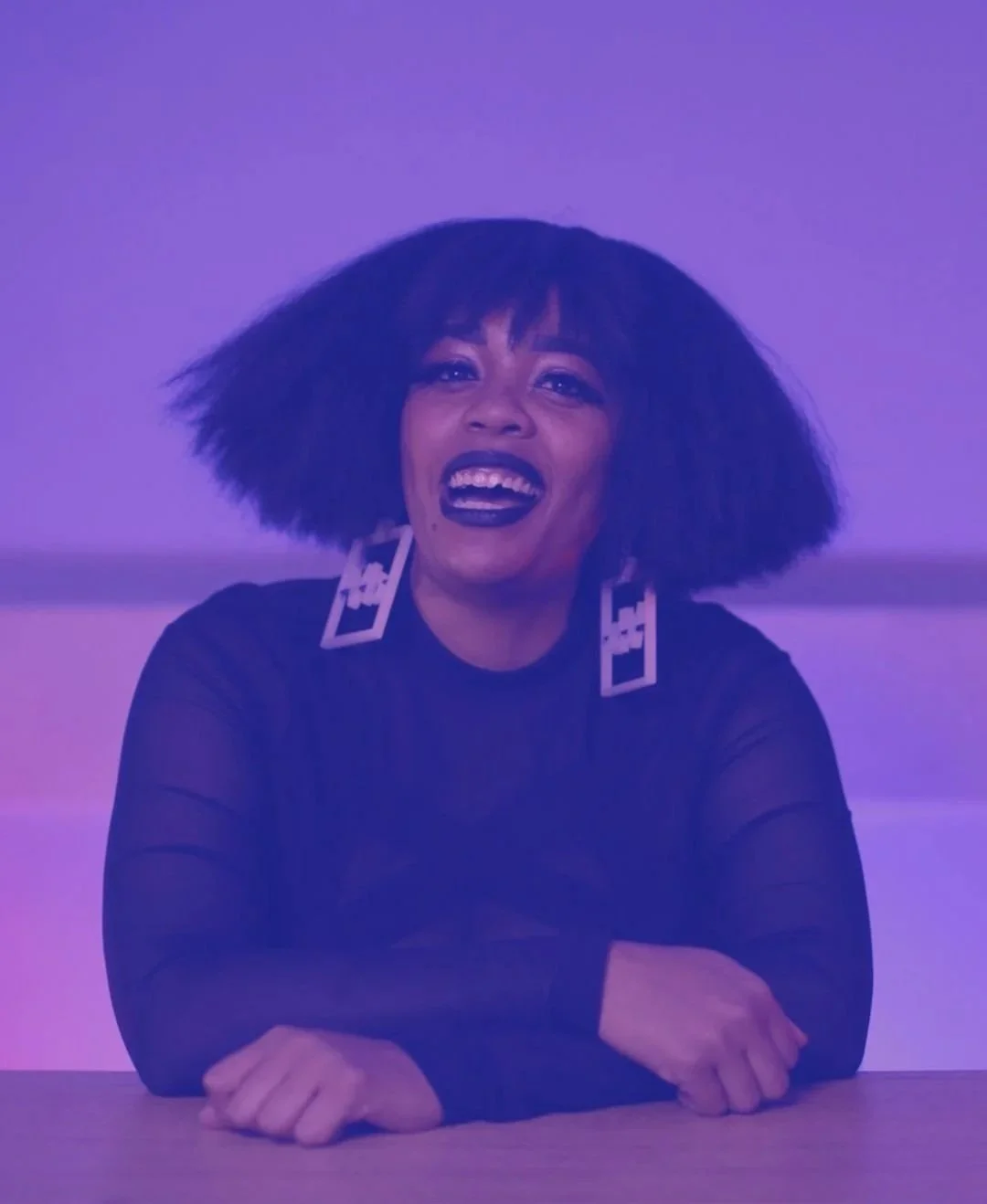 Woman with large curly hair and earrings, smiling with her mouth open, against a purple background.