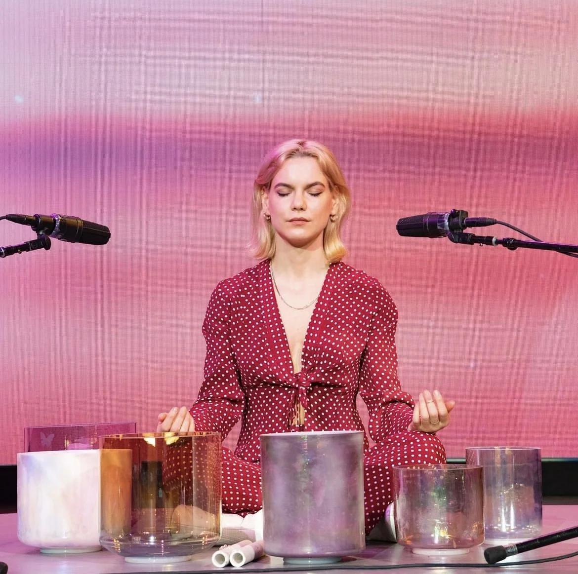 Woman sitting cross-legged with eyes closed, wearing a red polka dot outfit, meditating among singing bowls on a stage with pink background.