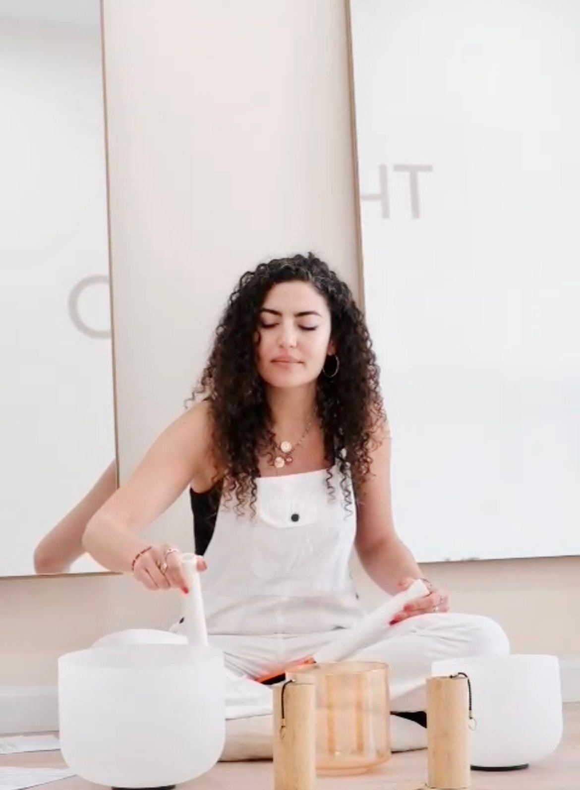 A young woman with curly hair and hoop earrings is sitting cross-legged on the floor, playing singing bowls with a mallet.