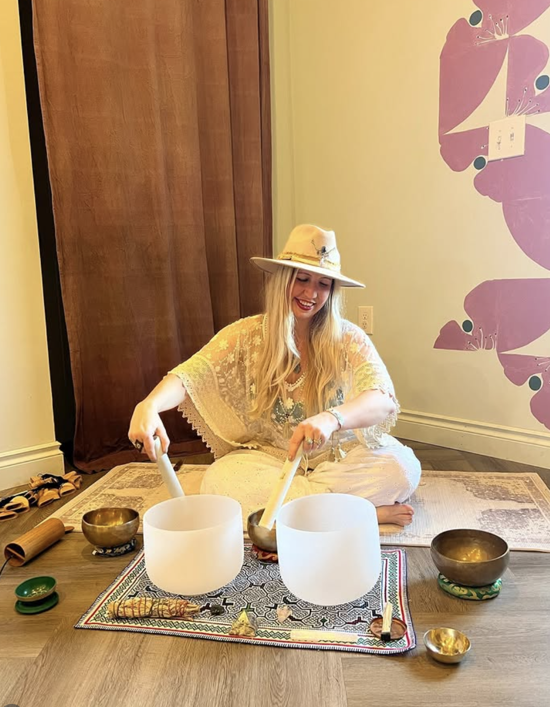 A woman wearing a wide-brimmed hat and bohemian clothing, sitting on the floor and playing singing bowls during a sound bath session in a room with pink floral wall art.