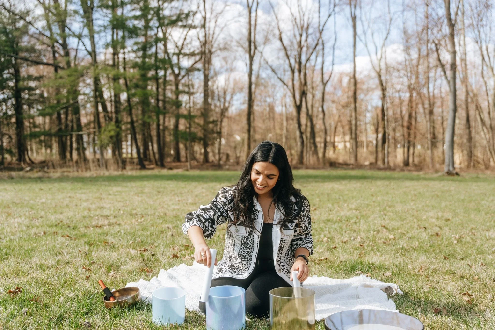 A woman smiling and sitting on a white blanket outdoors in a grassy field, playing hand chimes with various mallet instruments around her, with a background of trees and a partly cloudy sky.