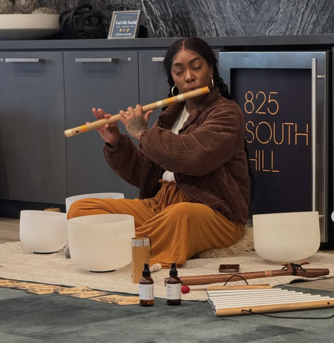 A woman sitting on a mat playing a wooden flute surrounded by white bowls, bottles, and a wooden stick, with a sign reading '825 South Hill' behind her.