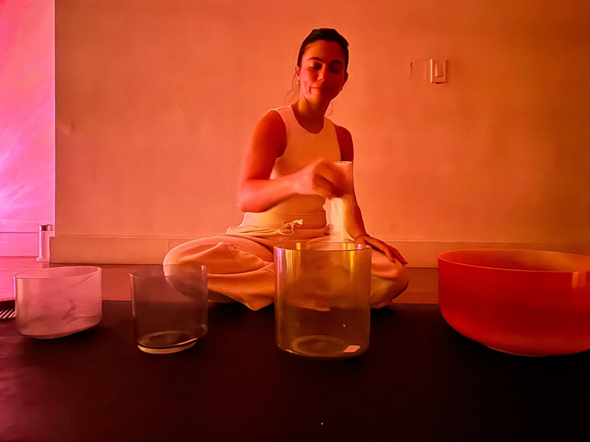 A woman sitting cross-legged on the floor, playing singing bowls of different sizes and colors in a dimly lit room.
