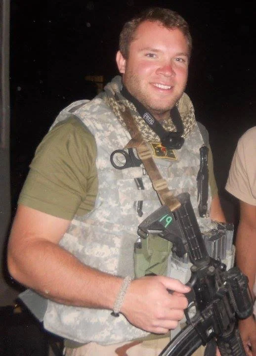 A man in military gear holding a rifle, smiling at the camera, standing in a dimly lit environment.