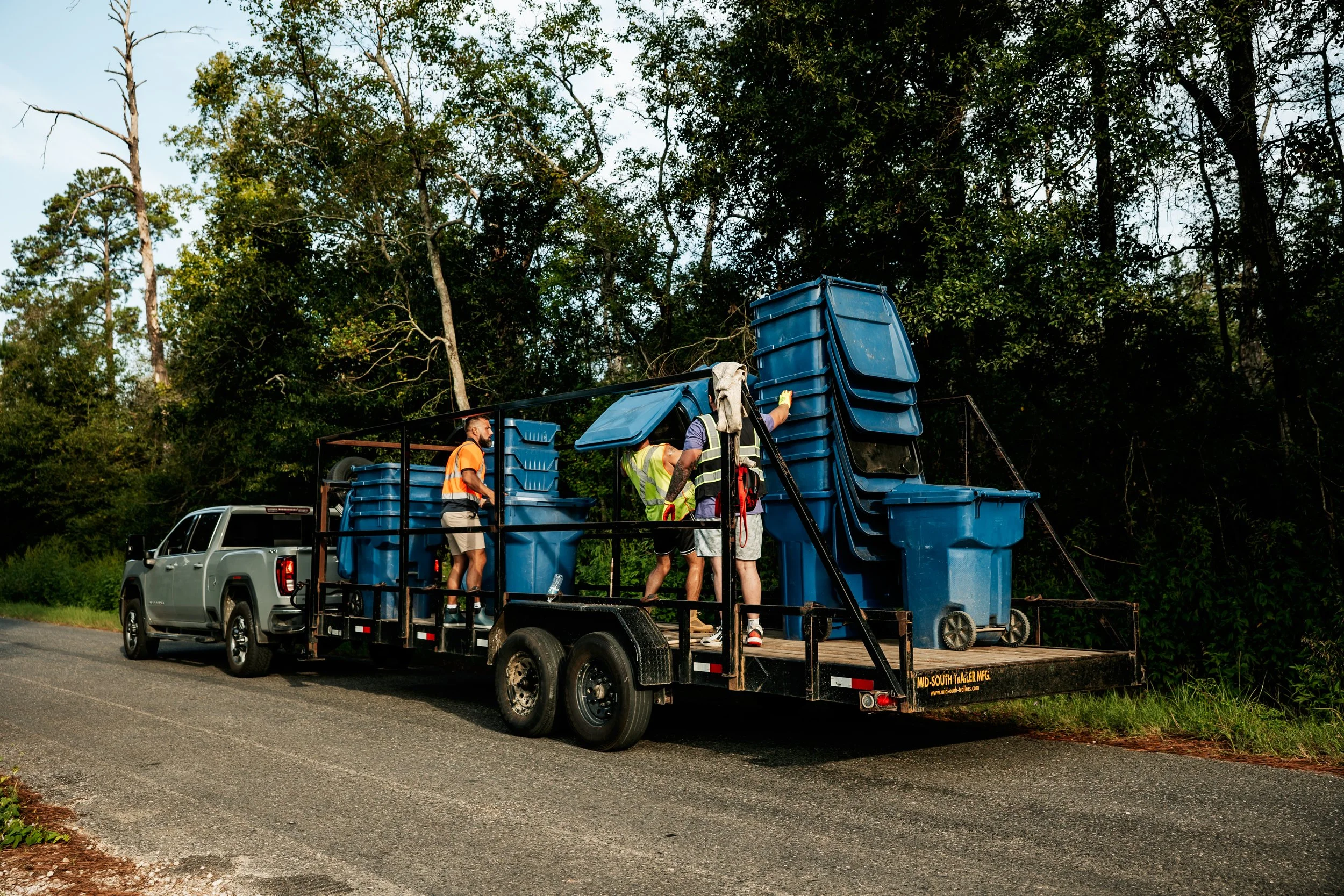 Workers loading blue trash bins onto a trailer attached to a pickup truck on the side of a road, with a wooded area in the background.