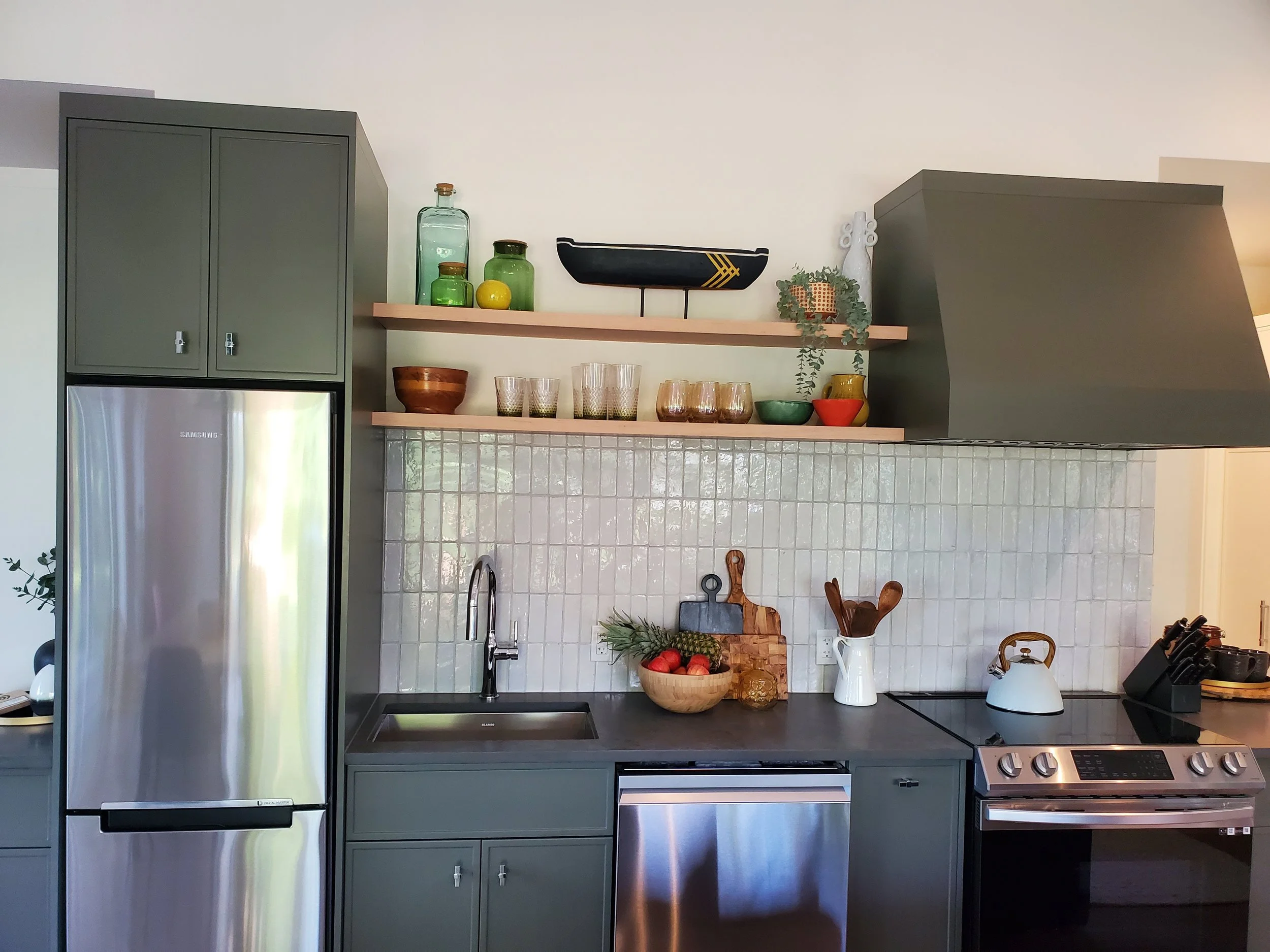 A modern kitchen with gray cabinets, open shelves with glass jars, bowls, and decorative items. Stainless steel refrigerator, sink, and stove are visible, along with kitchen utensils and a fruit bowl.