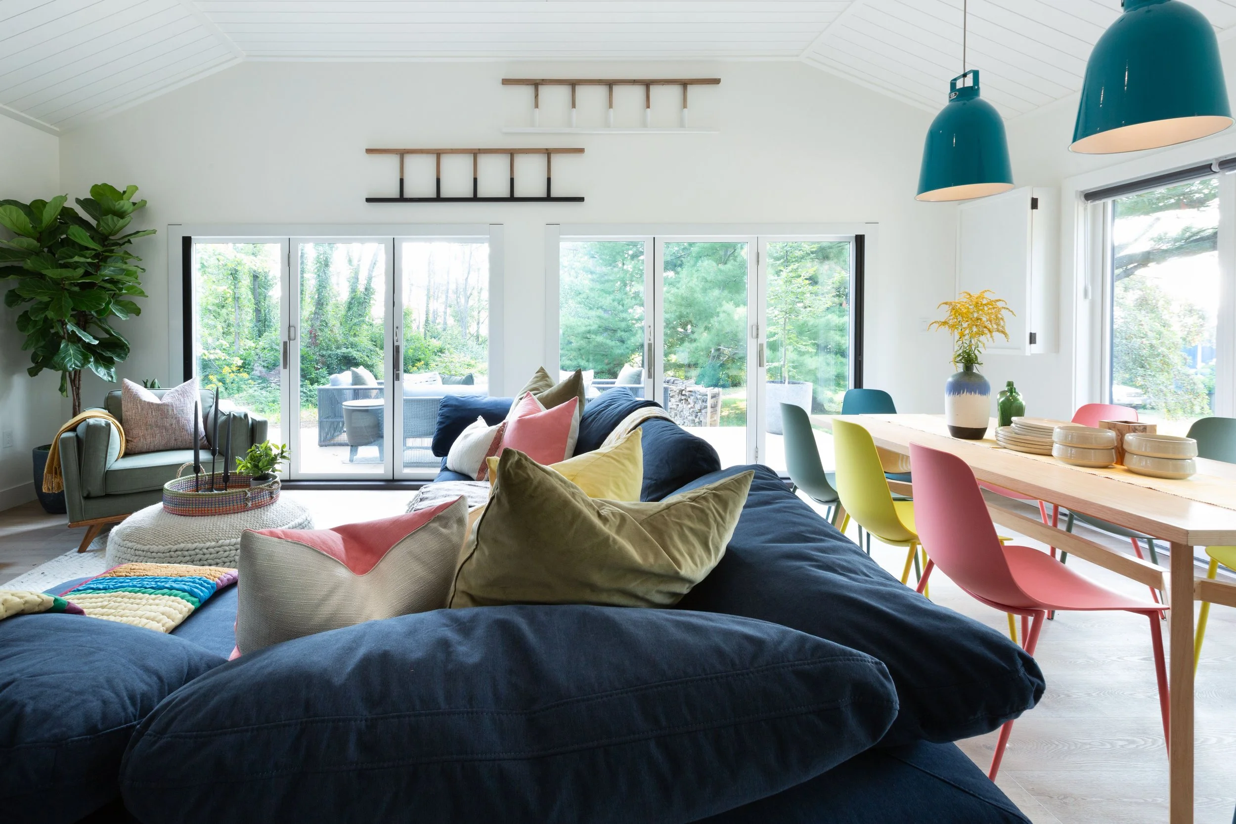 Bright living room with large windows showing green trees outside, featuring a navy blue sofa with multicolored pillows, a cream armchair with throw pillows, a wooden dining table with colorful chairs, and pendant lights hanging from the ceiling.