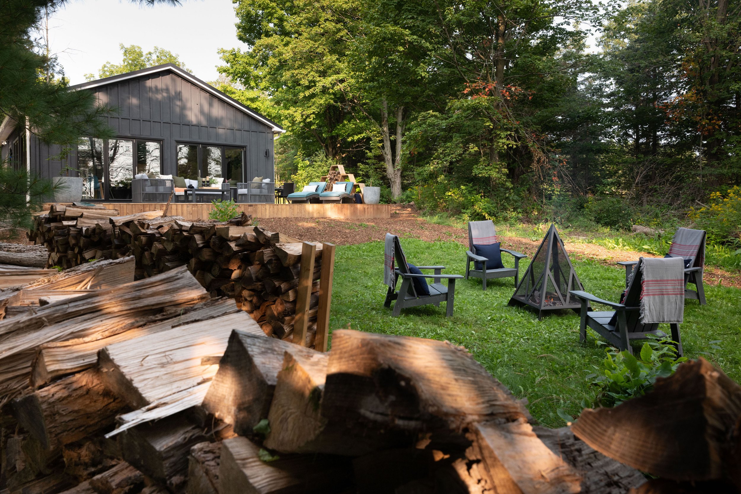 A backyard with a fire pit, outdoor chairs with throws, a log cabin-style house with a deck, and a pile of firewood, surrounded by trees and greenery.
