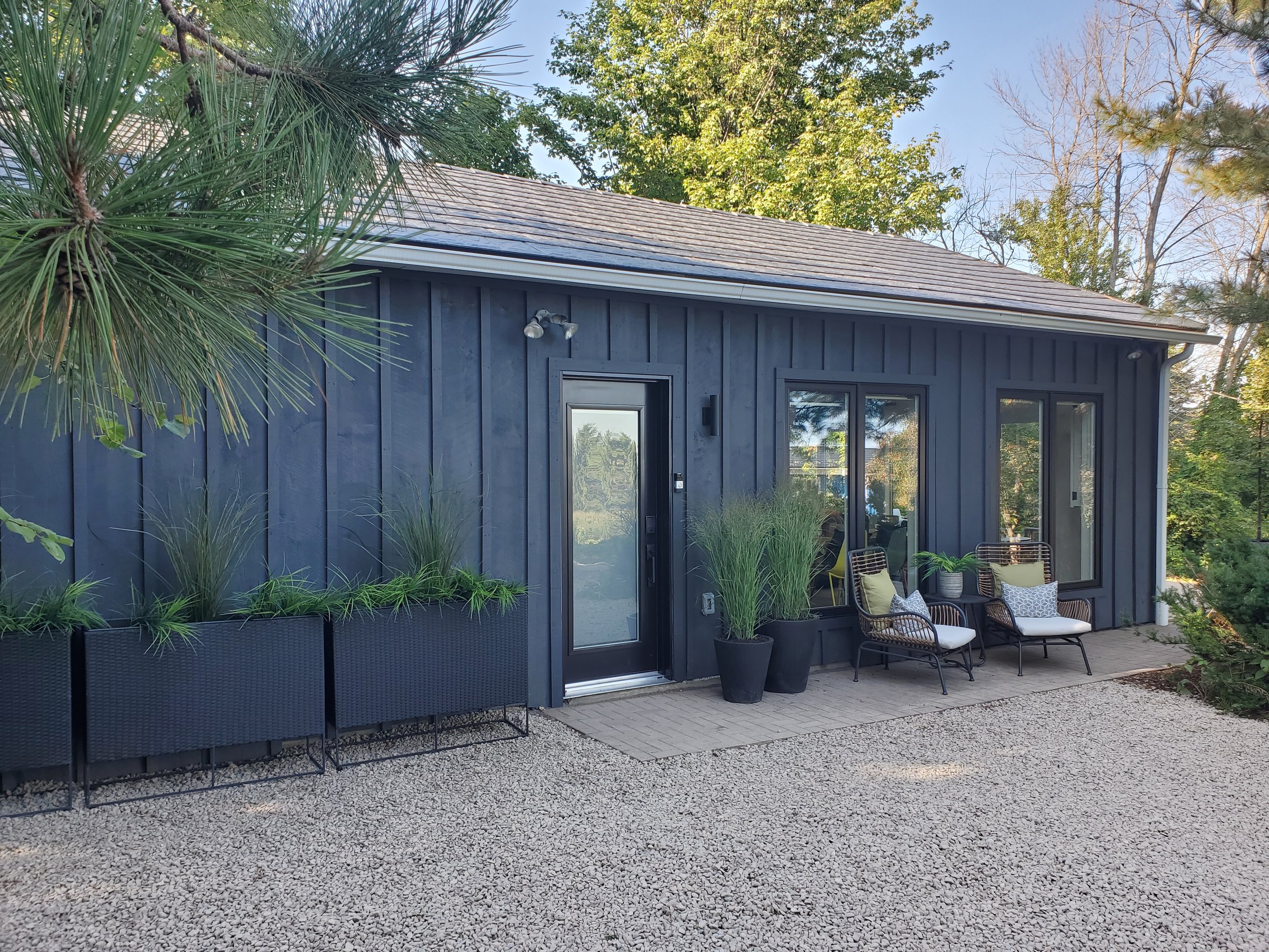 A modern house exterior with navy blue vertical siding, large glass doors, and outdoor sitting area with two chairs and a cushion, surrounded by greenery and trees.