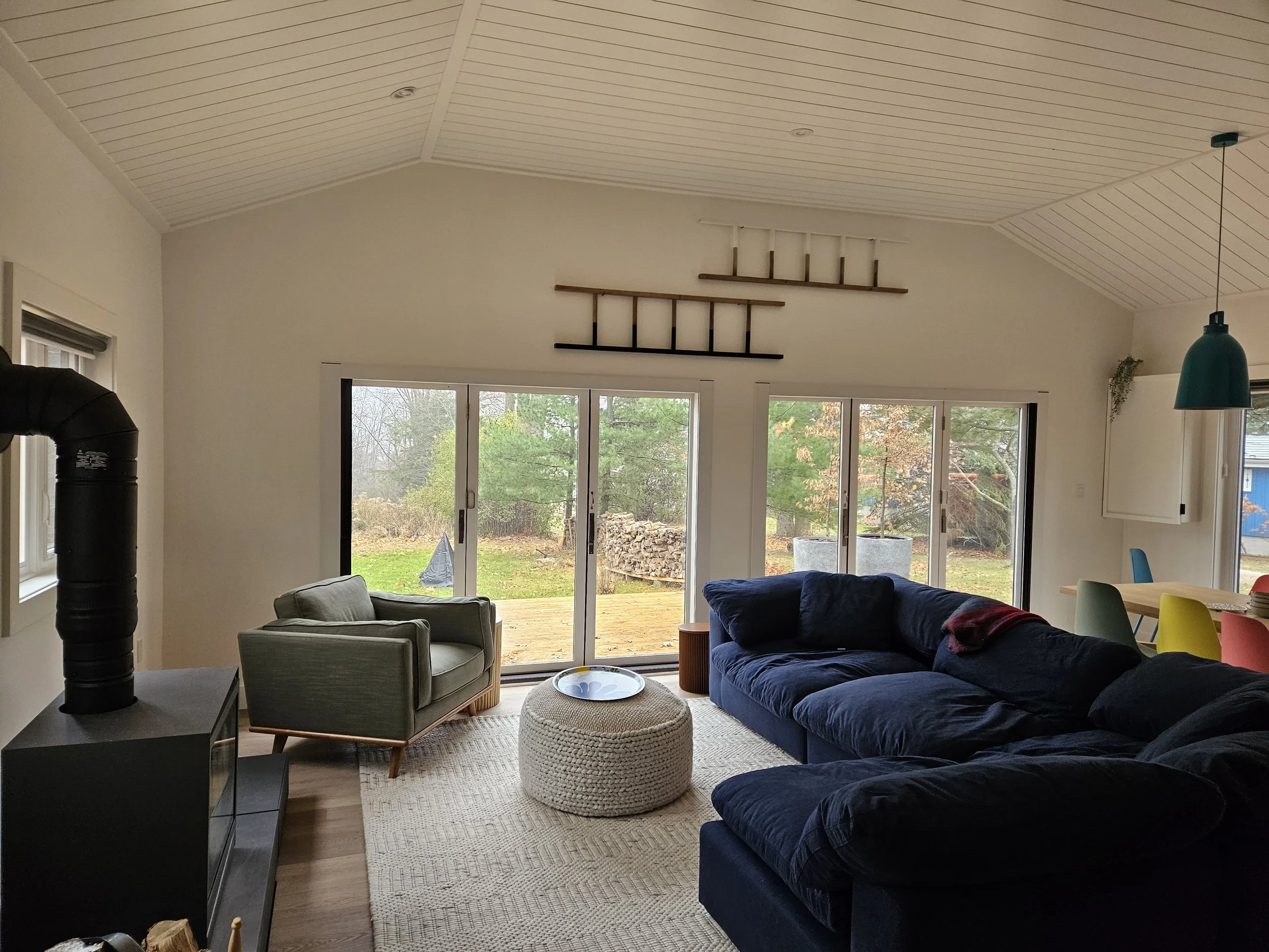A living room with a vaulted, white-shiplap ceiling, large windows with sliding doors showing an outside yard with trees and stacked firewood, and a seating area with a navy couch, a green armchair, and a white woven ottoman.