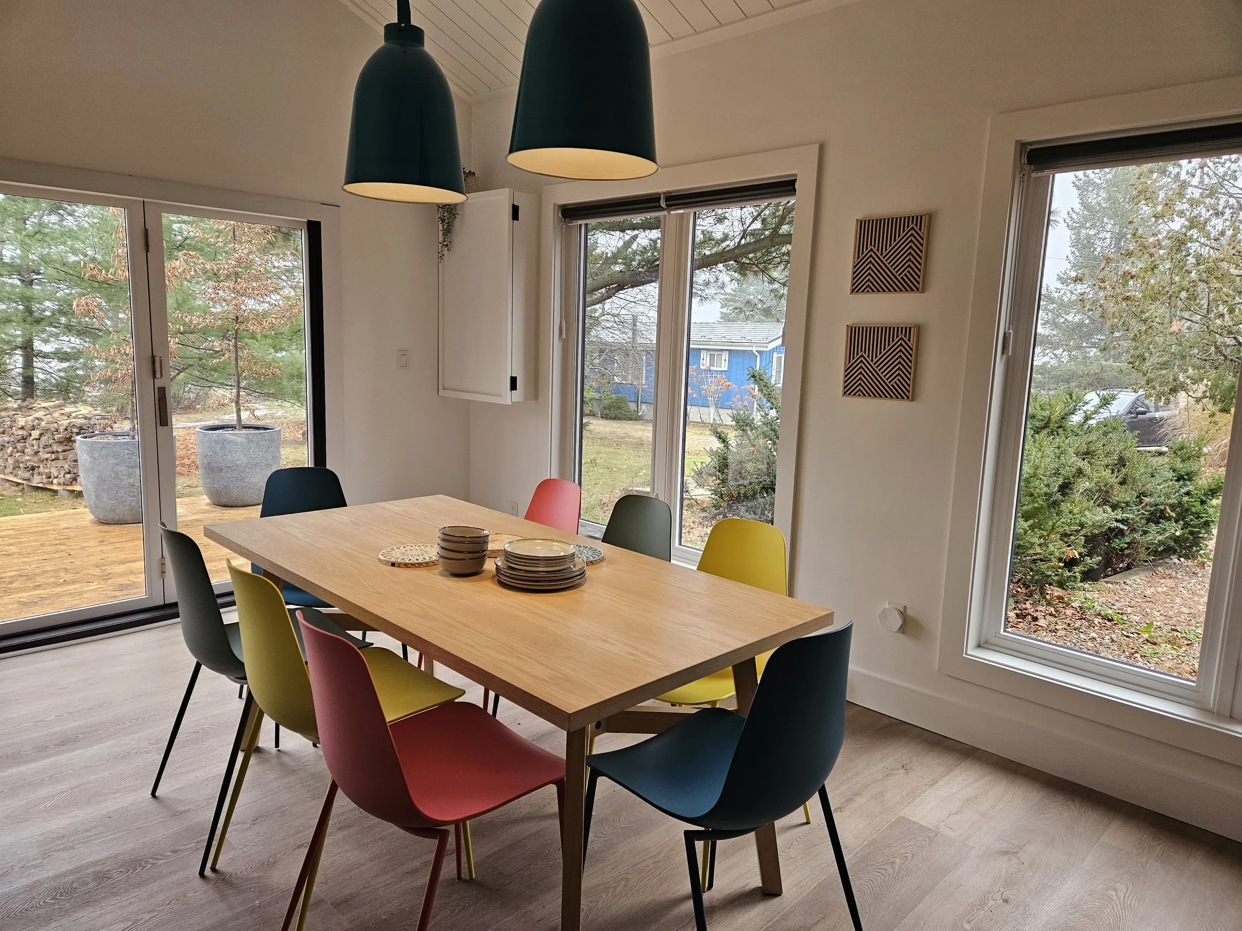 A dining room with a wooden table and colorful chairs, large windows with views of trees and houses outside, and modern black pendant lights hanging from the ceiling.