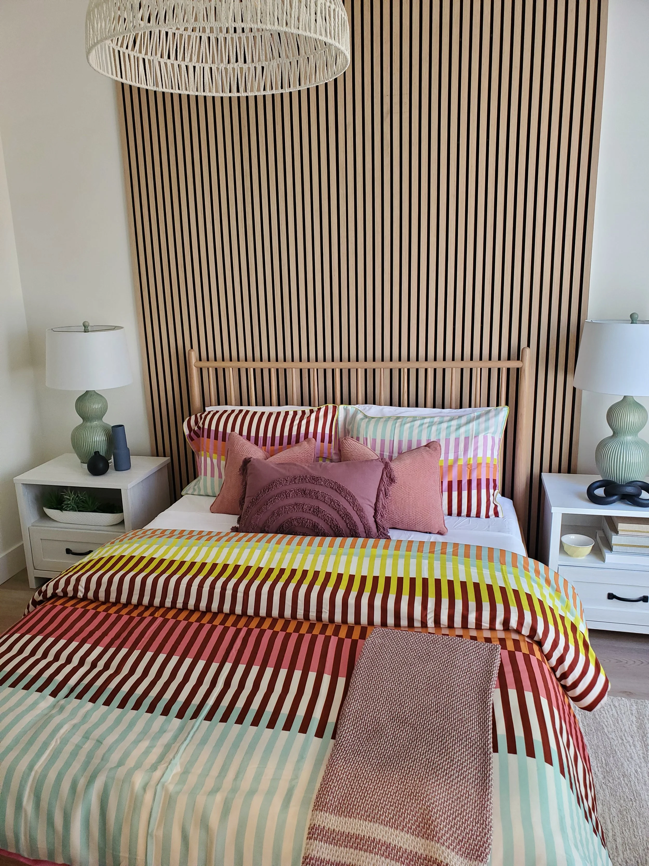 A neatly made bed with colorful striped bedding, pillows, and a throw blanket, flanked by two white nightstands with green lamps on each side, against a wooden slat accent wall.
